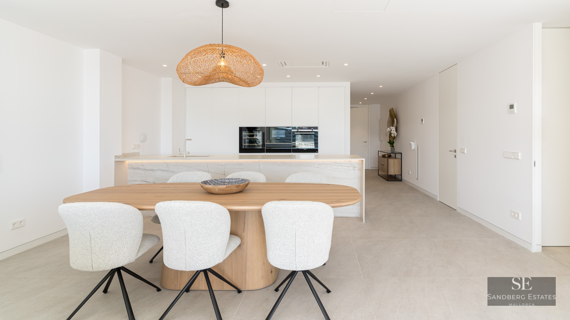 A bright dining area with a wooden table, white chairs, and a wicker pendant light in front of a modern kitchen.