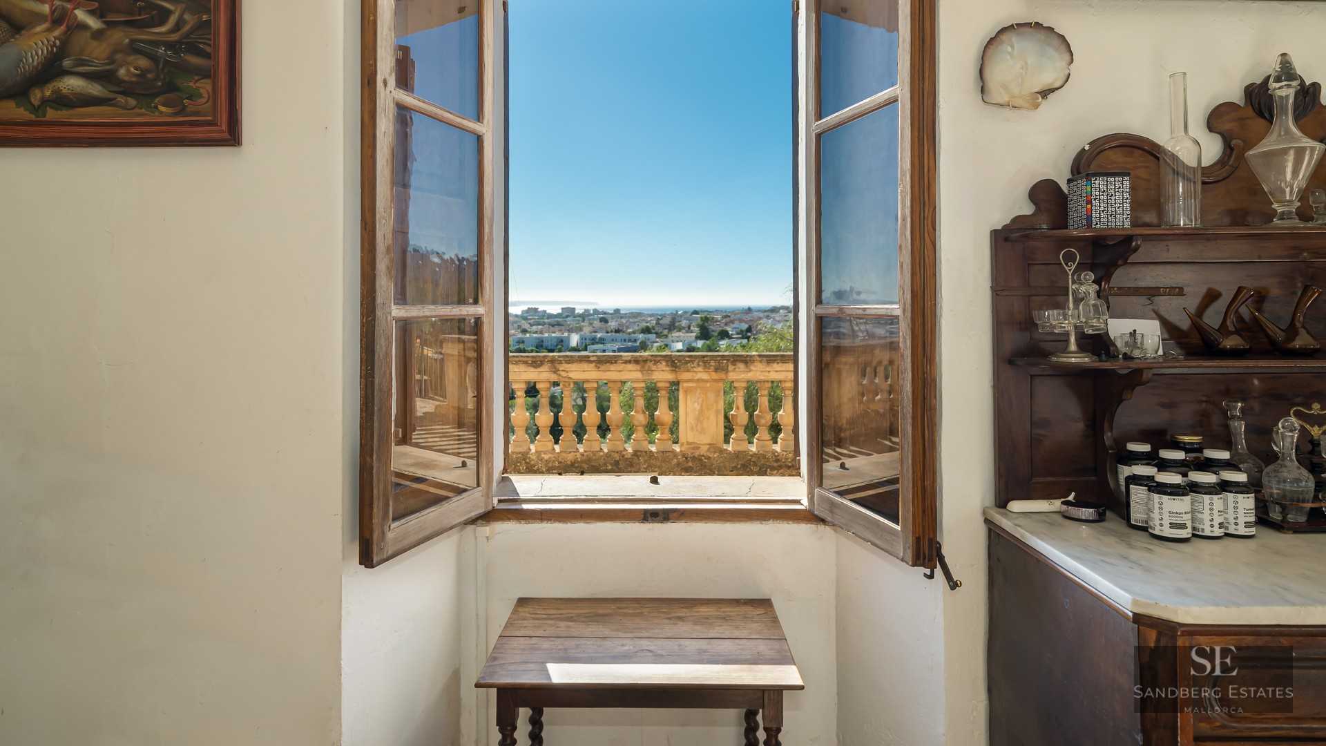 An open wooden window framing a panoramic view of a coastal city and the sea under a clear blue sky.