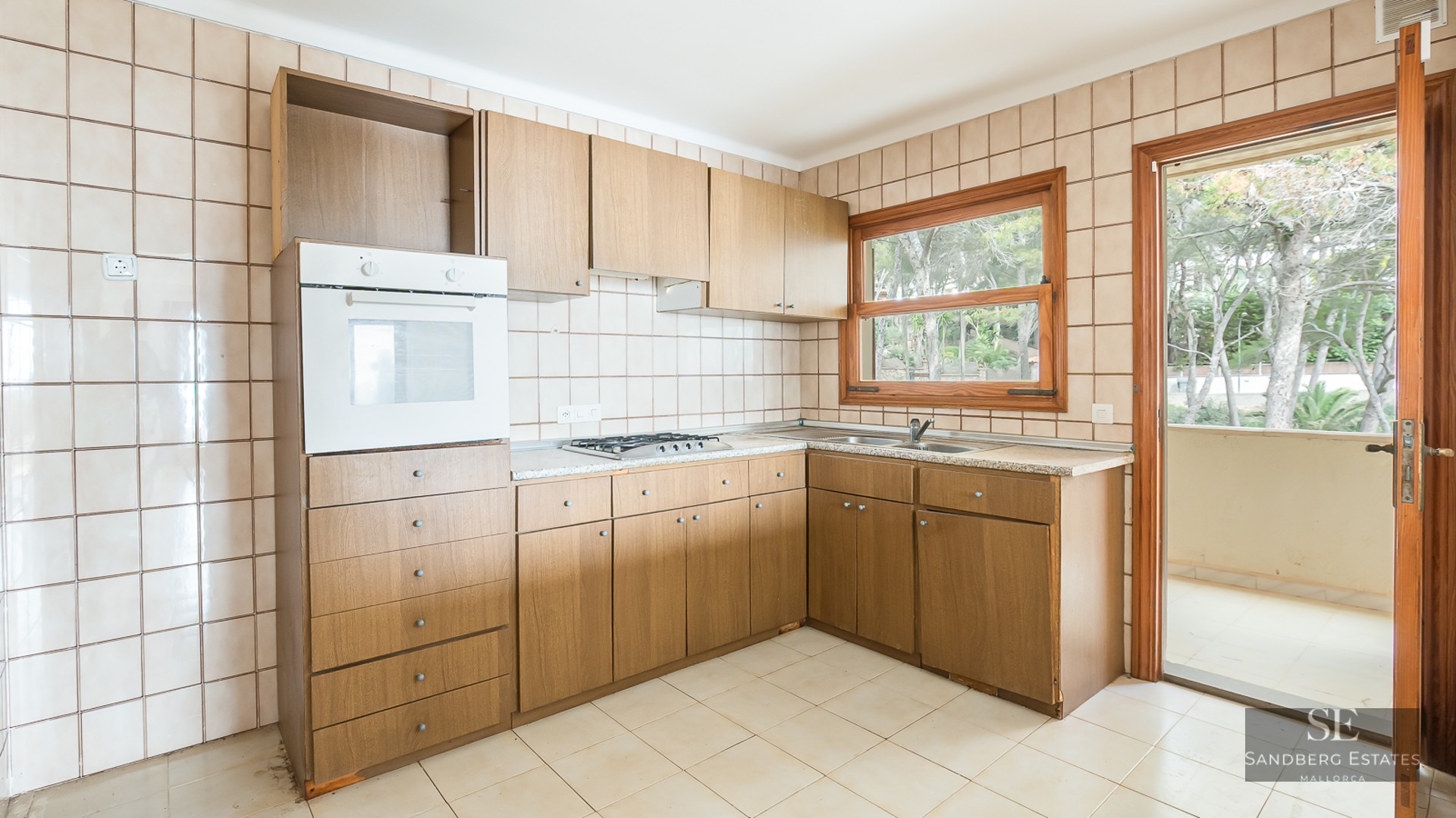 Bright kitchen featuring wooden cabinetry, beige wall tiles, and views of greenery through a window and door.
