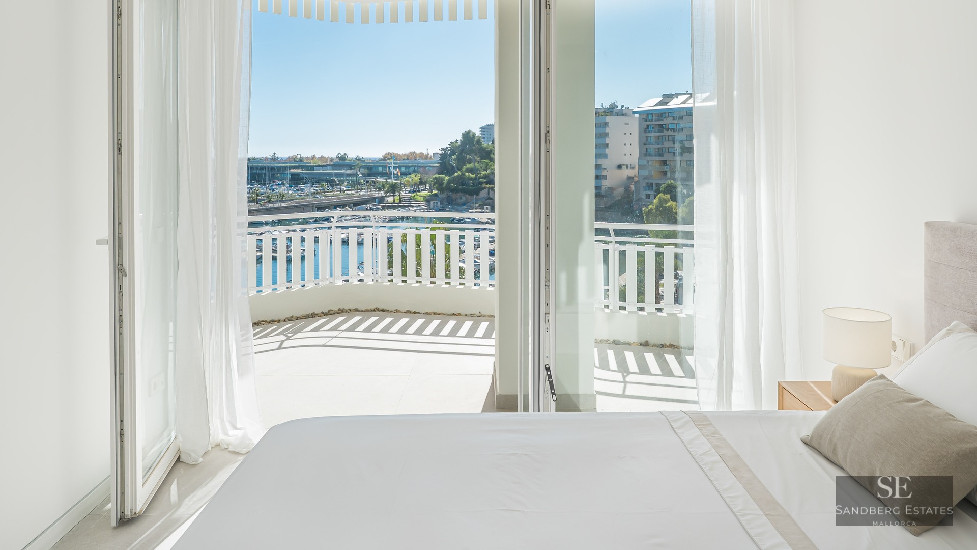Bright bedroom with white bed and open glass doors leading to a balcony overlooking a marina with boats.