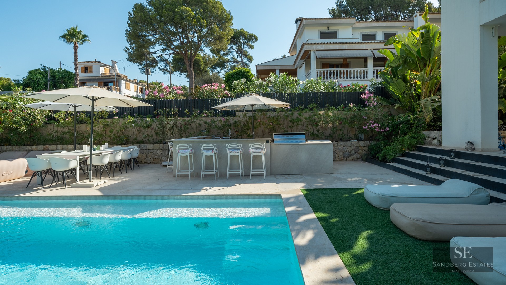 Bright swimming pool area with modern outdoor kitchen, bar stools, and dining set under umbrellas in a sunny garden.
