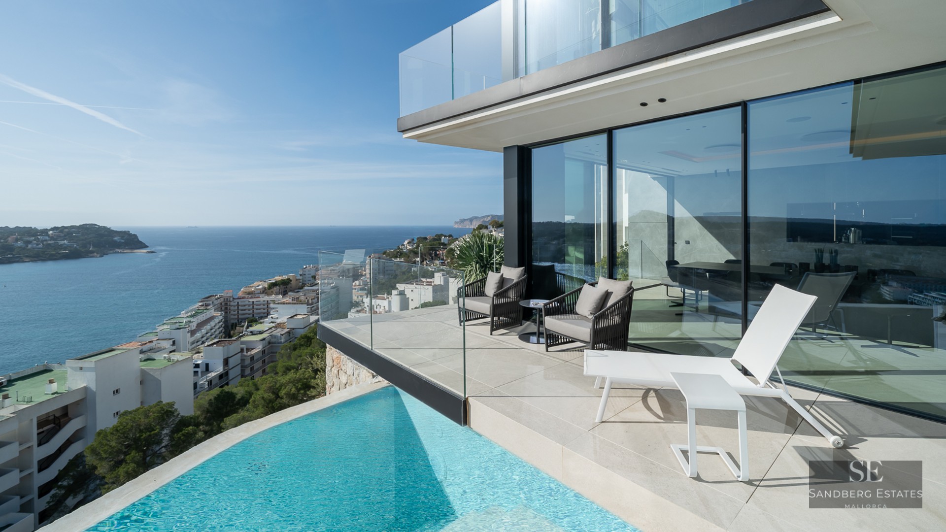 Modern terrace featuring a turquoise infinity pool, white sun lounger, and glass railings overlooking the Mediterranean sea.