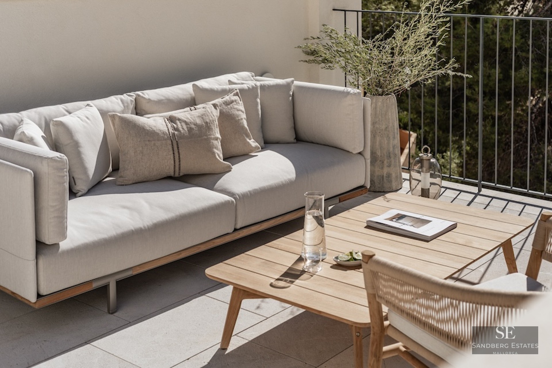 Modern outdoor terrace featuring a plush grey sofa, wooden coffee table, and greenery against a white wall and railing.