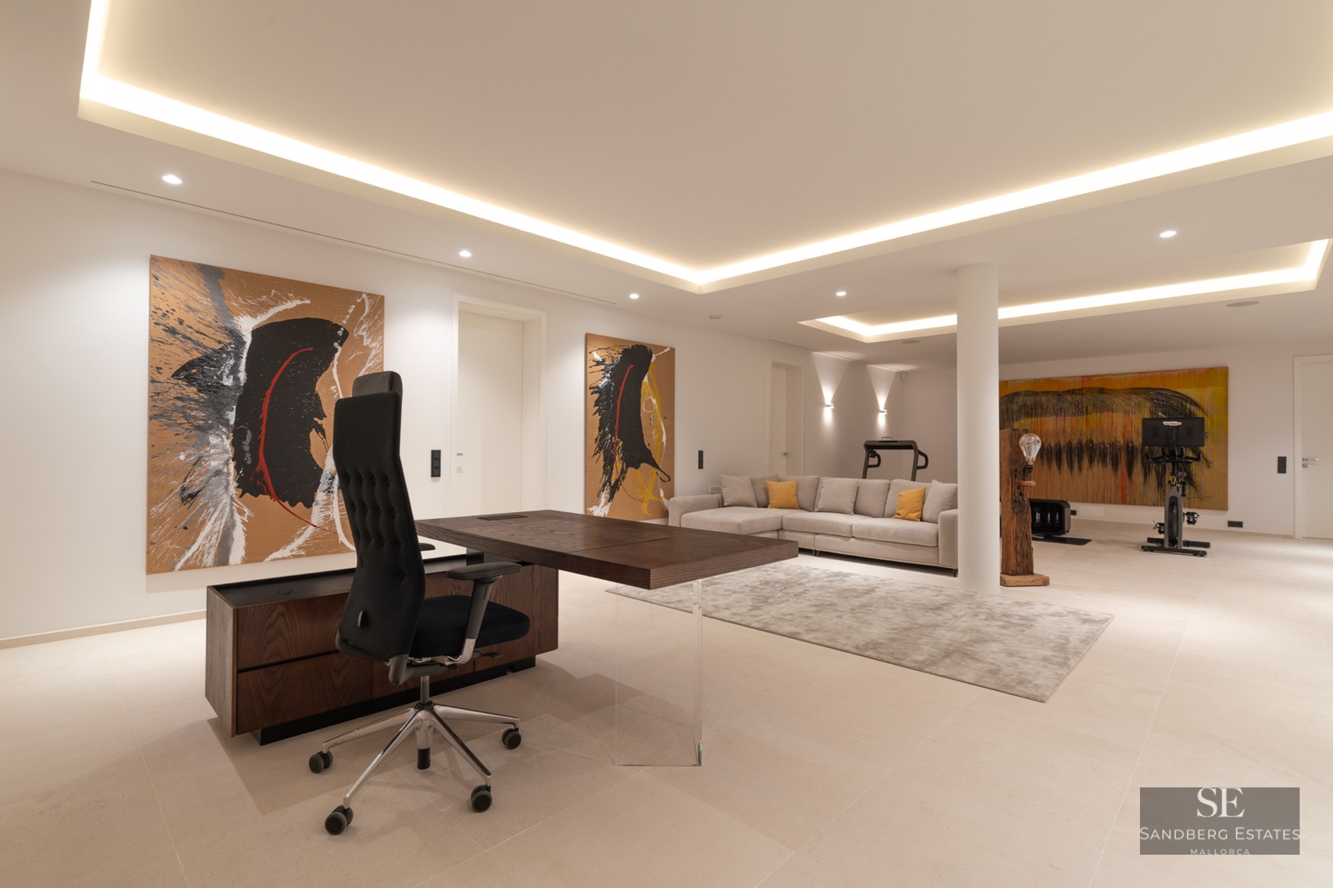 Spacious modern home office with a dark wood desk, grey sofa, abstract art, and gym equipment in the background.