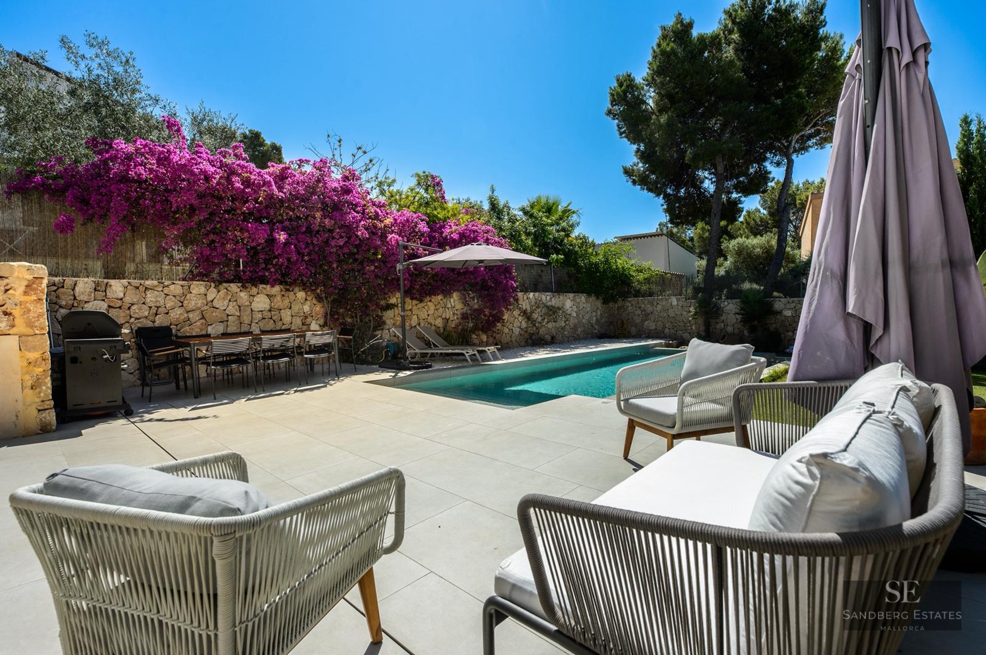 Modern private pool area with outdoor lounge seating, dining table, BBQ, and vibrant purple bougainvillea on a stone wall.
