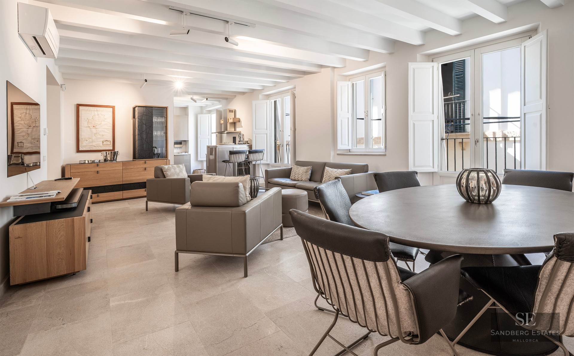 Salon spacieux avec plafonds à poutres blanches, meubles en cuir gris et table à manger ronde.