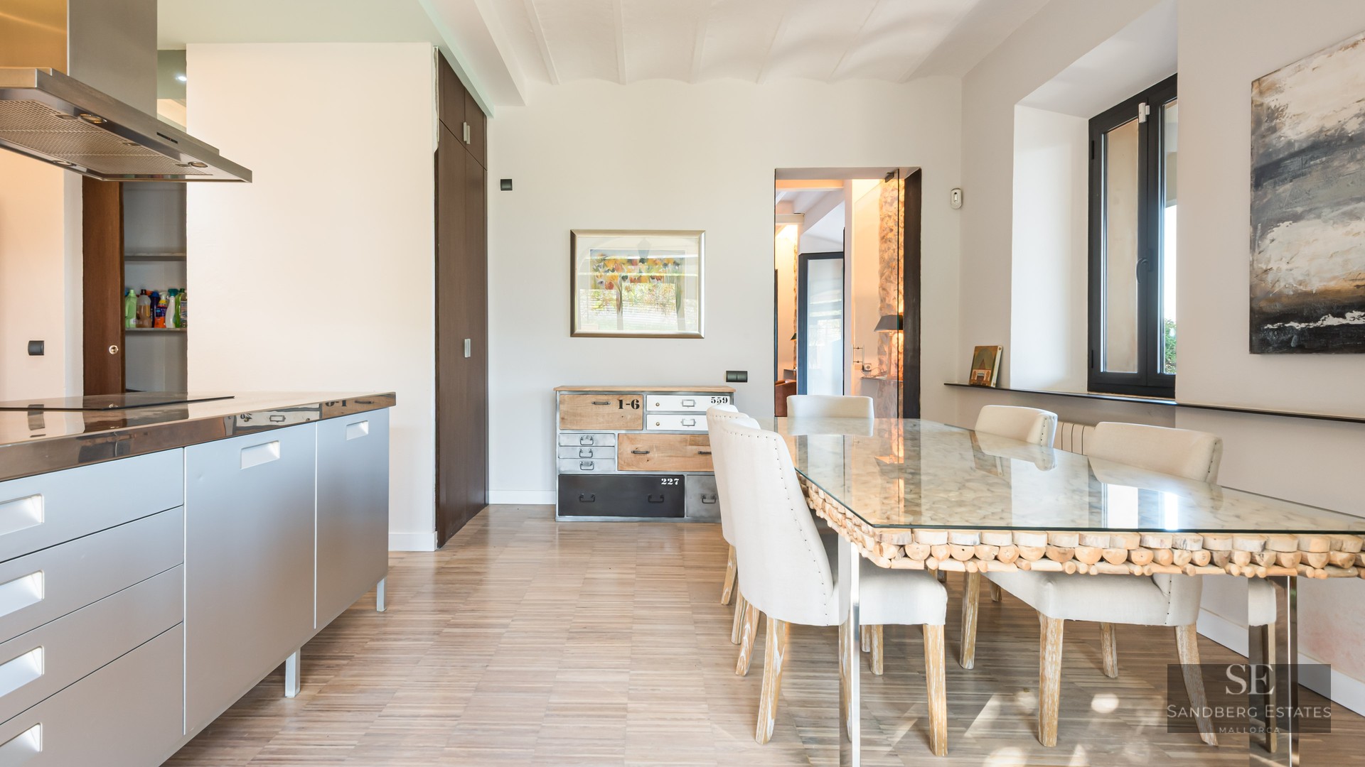 Bright dining room featuring a glass-top table with wood base, white chairs, and a stainless steel kitchen unit.