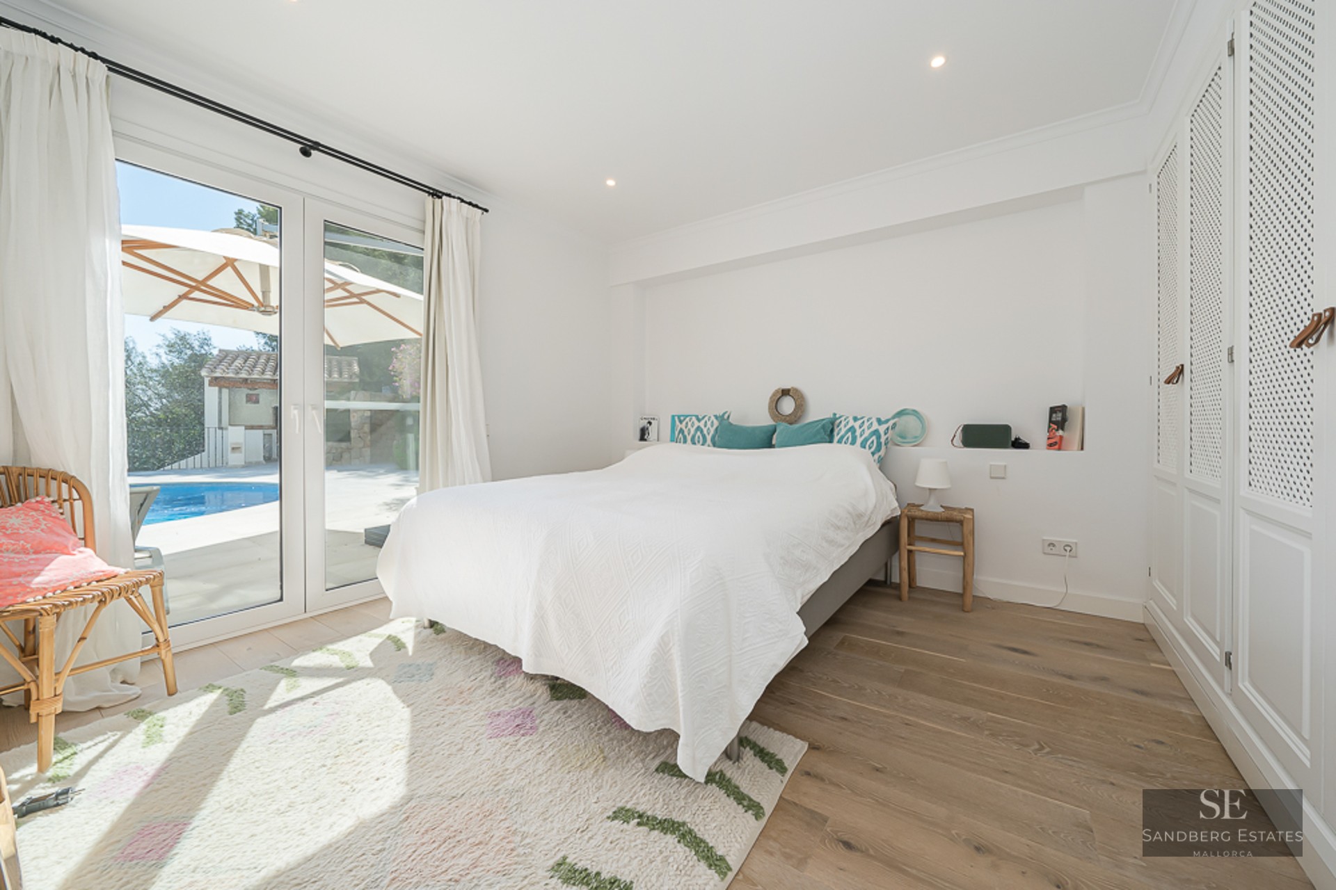 Chambre lumineuse avec un grand lit, parquet et portes vitrées s'ouvrant sur une piscine turquoise et un parasol.