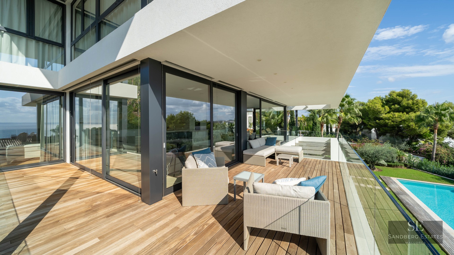 Spacious wooden terrace with designer lounge furniture and glass railings overlooking a swimming pool and lush garden.