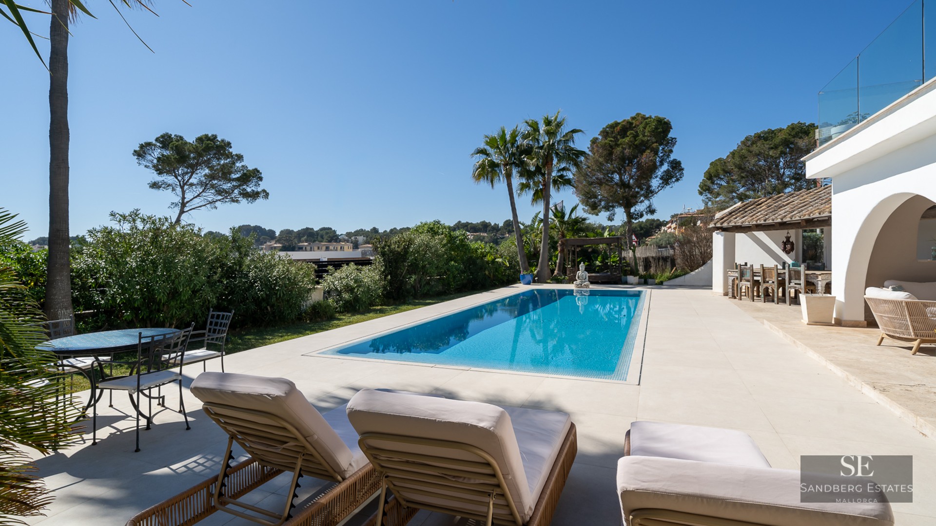 Turquoise swimming pool surrounded by white stone tiles, sun loungers, palm trees, and a Mediterranean villa under a blue sky.