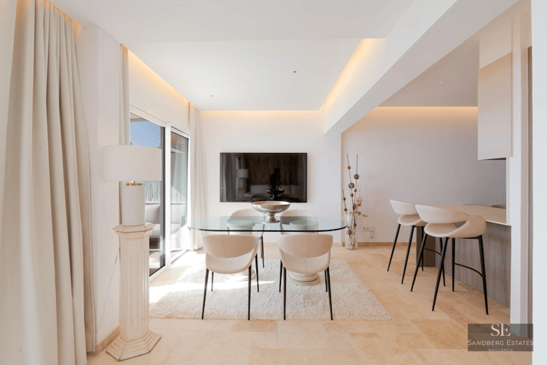 Minimalist dining room featuring a glass table, cream chairs, white rug, and a breakfast bar in the background.
