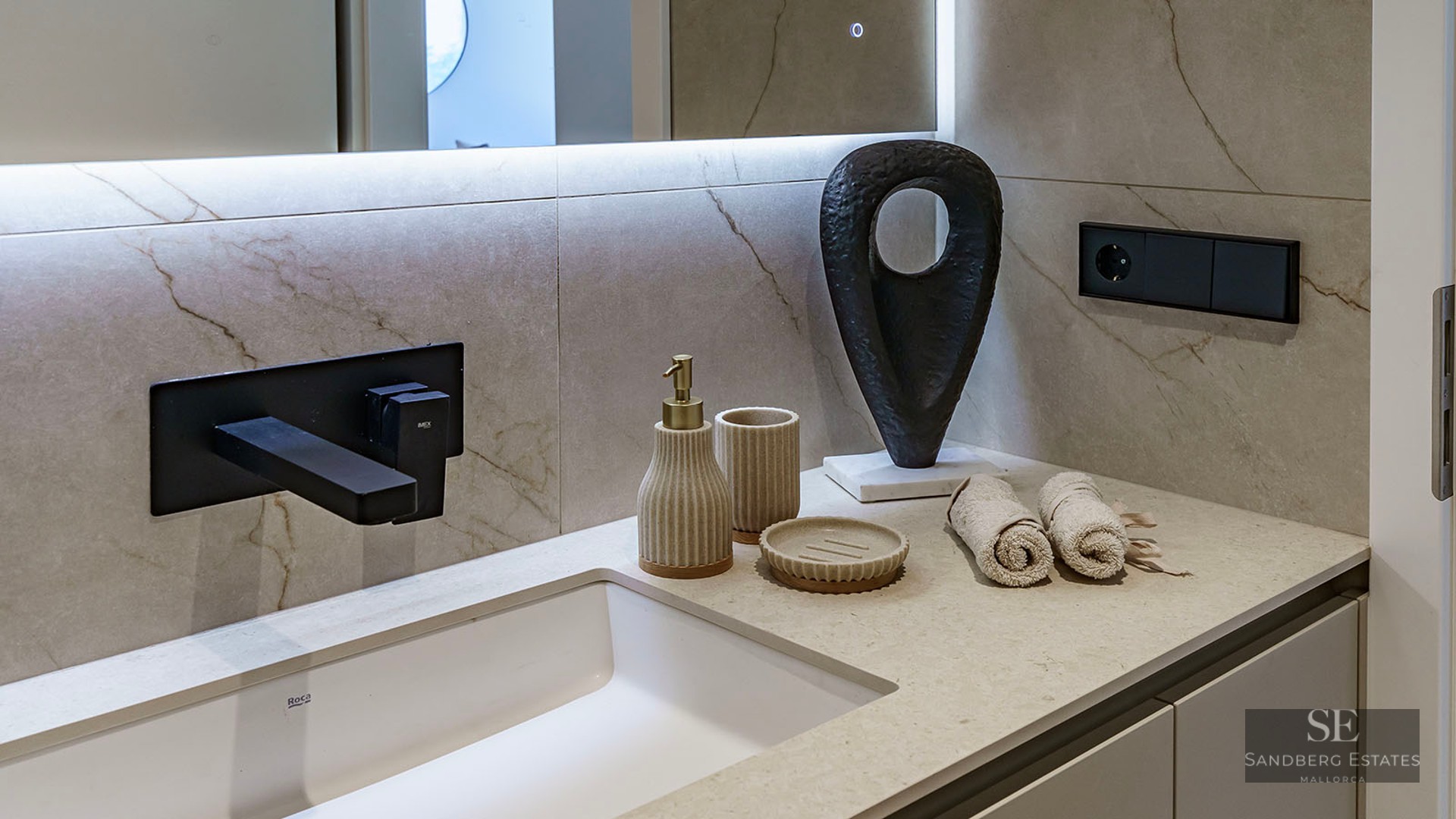 Close-up of a white sink with a matte black wall-mounted faucet, beige marble-effect tiling, and designer accessories.