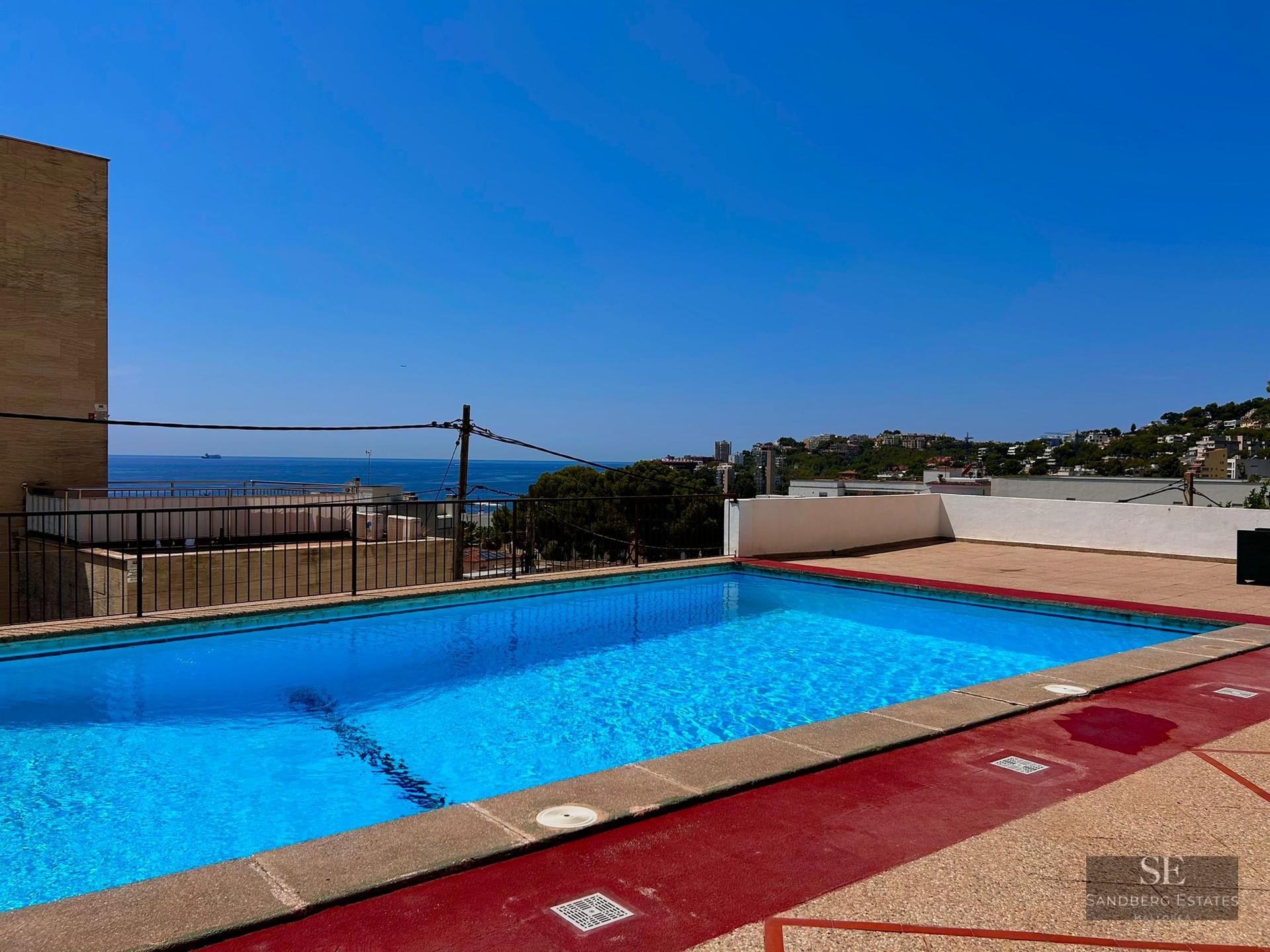 Blue rooftop pool with red deck overlooking the Mediterranean sea and coastline under a clear blue sky.