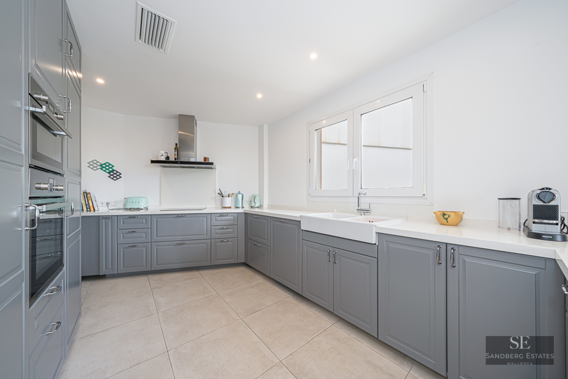 Cuisine lumineuse avec placards gris style shaker, plans de travail blancs et grand évier timbre d'office.