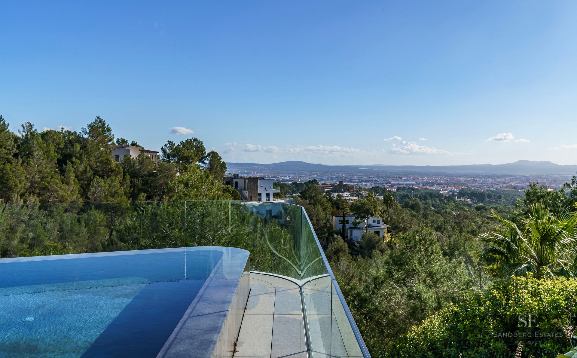 An infinity-edge pool with glass railing overlooking a lush forested valley and distant city.