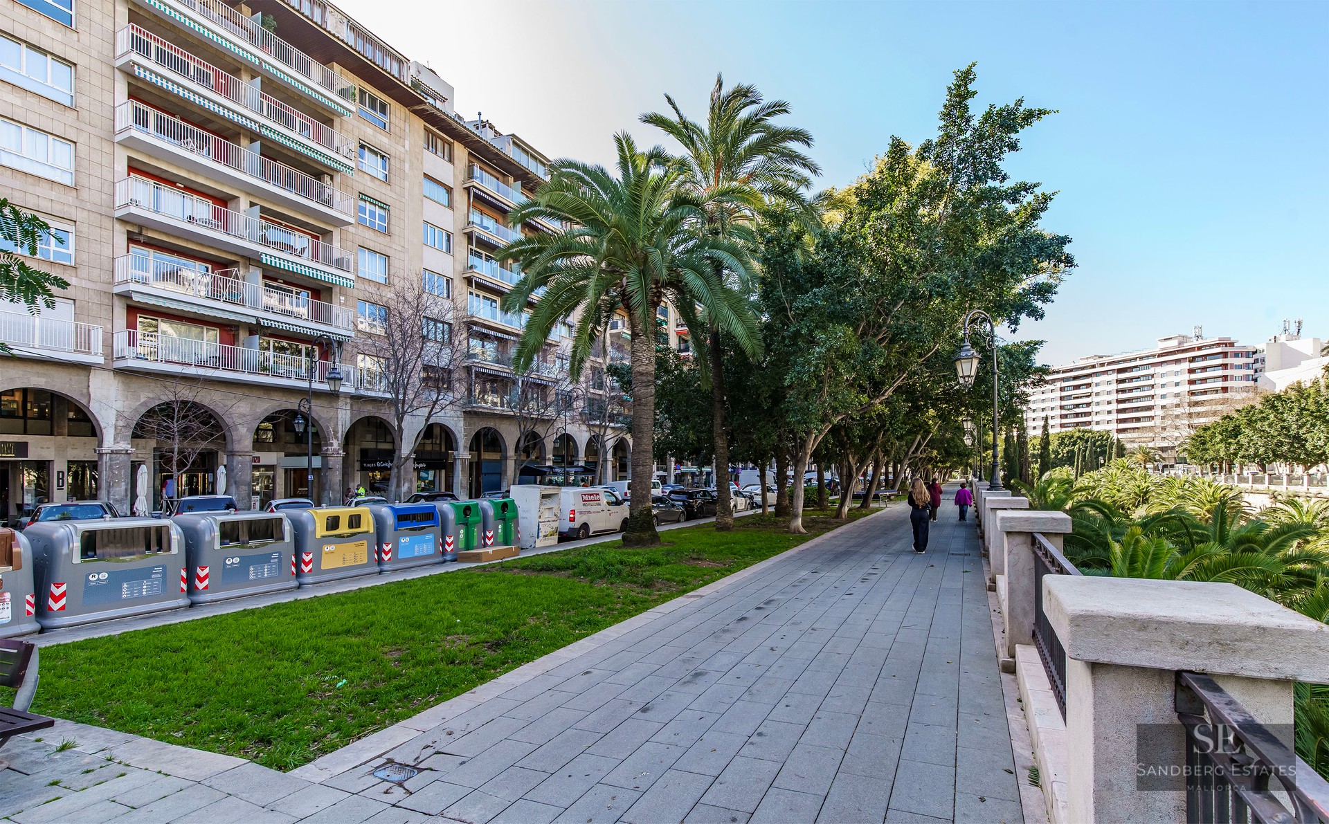 Promenade urbaine pavée avec palmiers à côté d'un immeuble en pierre à plusieurs étages avec balcons sous un ciel bleu.