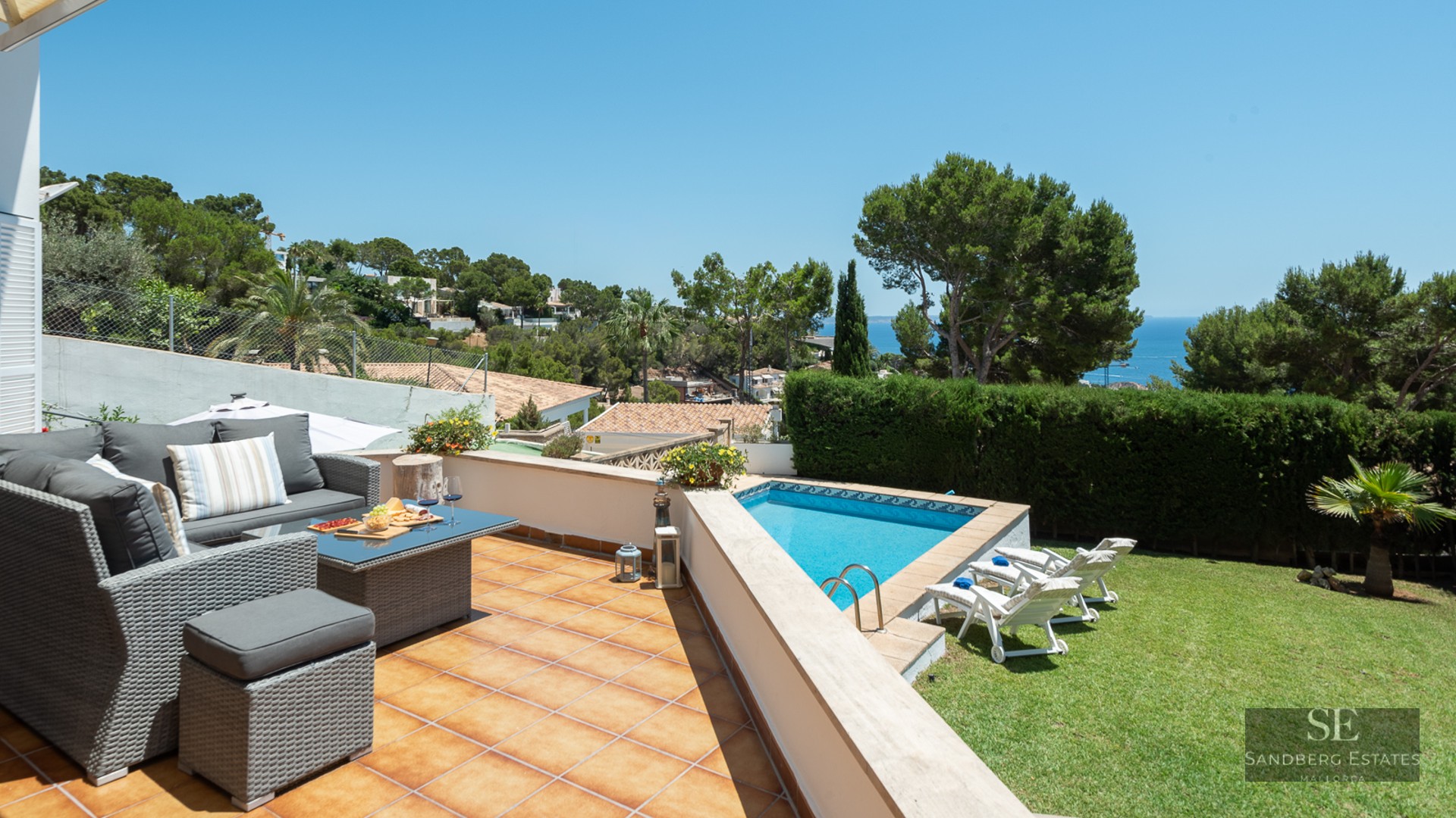 Sun-drenched terrace with grey wicker sofas, a blue swimming pool, green lawn, and panoramic ocean views.