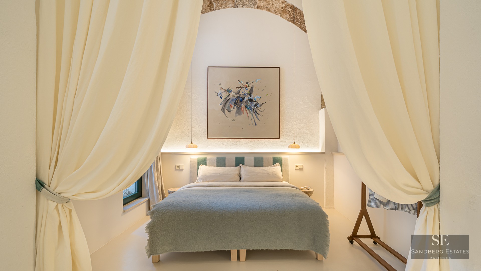 Chambre élégante avec grand lit, rideaux crème, arche en pierre apparente et tableau abstrait moderne.