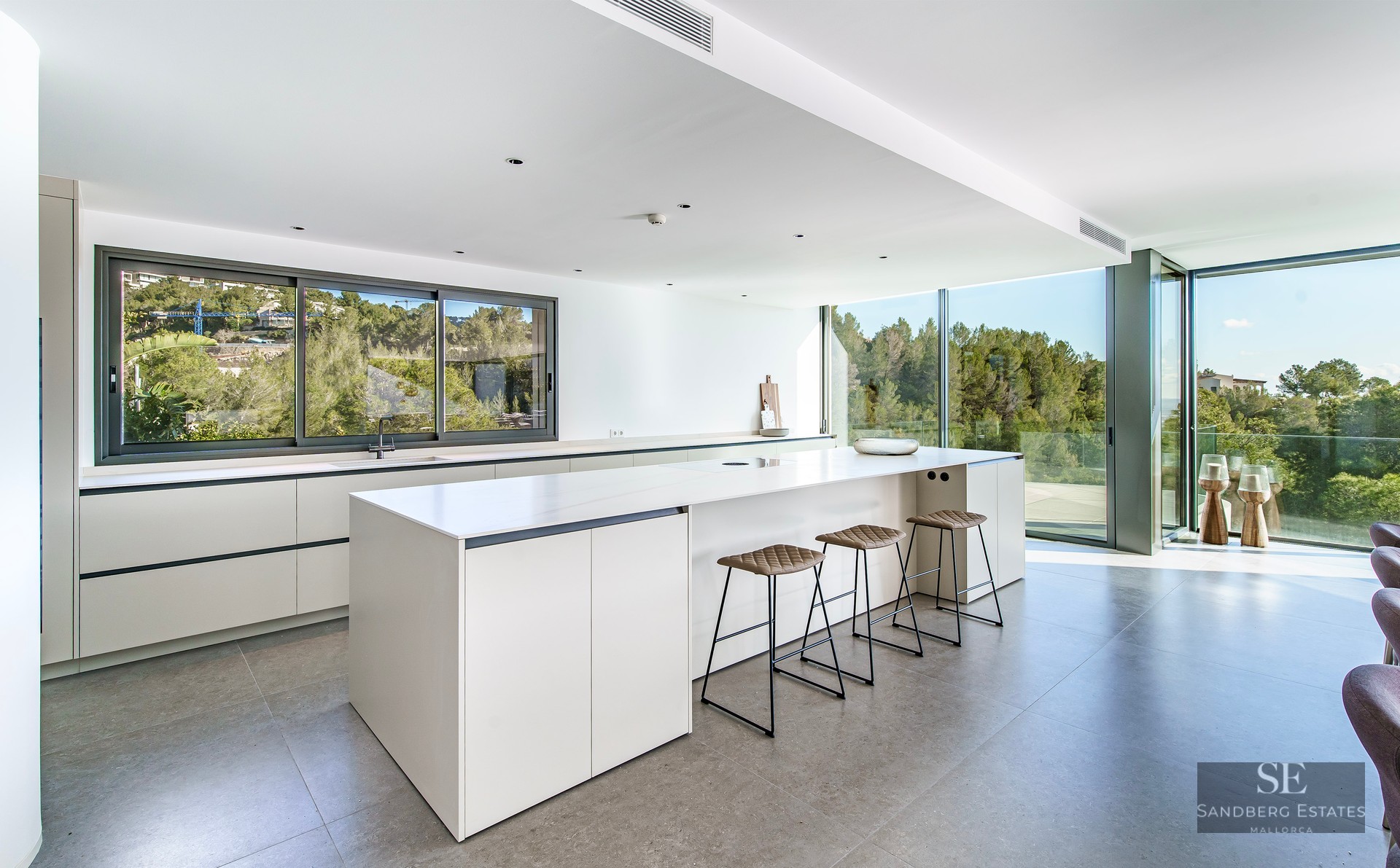 Bright kitchen featuring a large white island, leather stools, and floor-to-ceiling windows overlooking a forest.