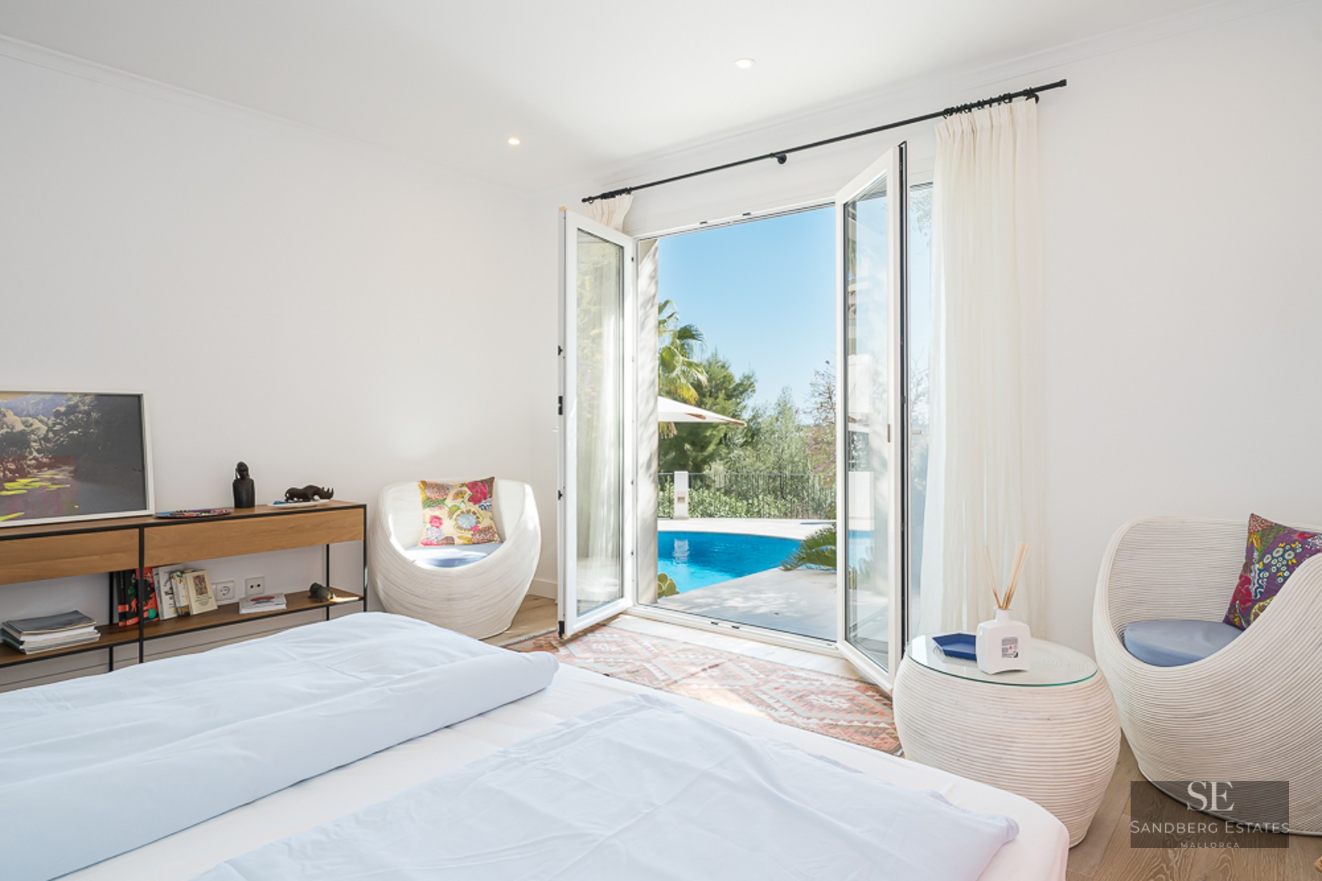Chambre lumineuse avec lit blanc et chaises design, s'ouvrant sur une piscine turquoise et un jardin.
