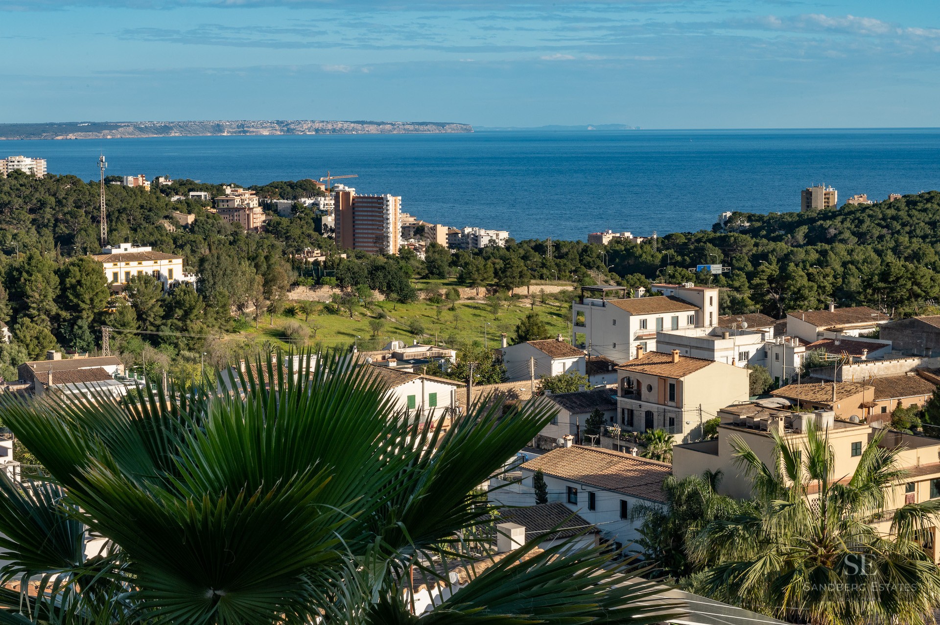 Elevated view of a coastal town with Mediterranean houses, lush greenery, and the blue sea in the distance.