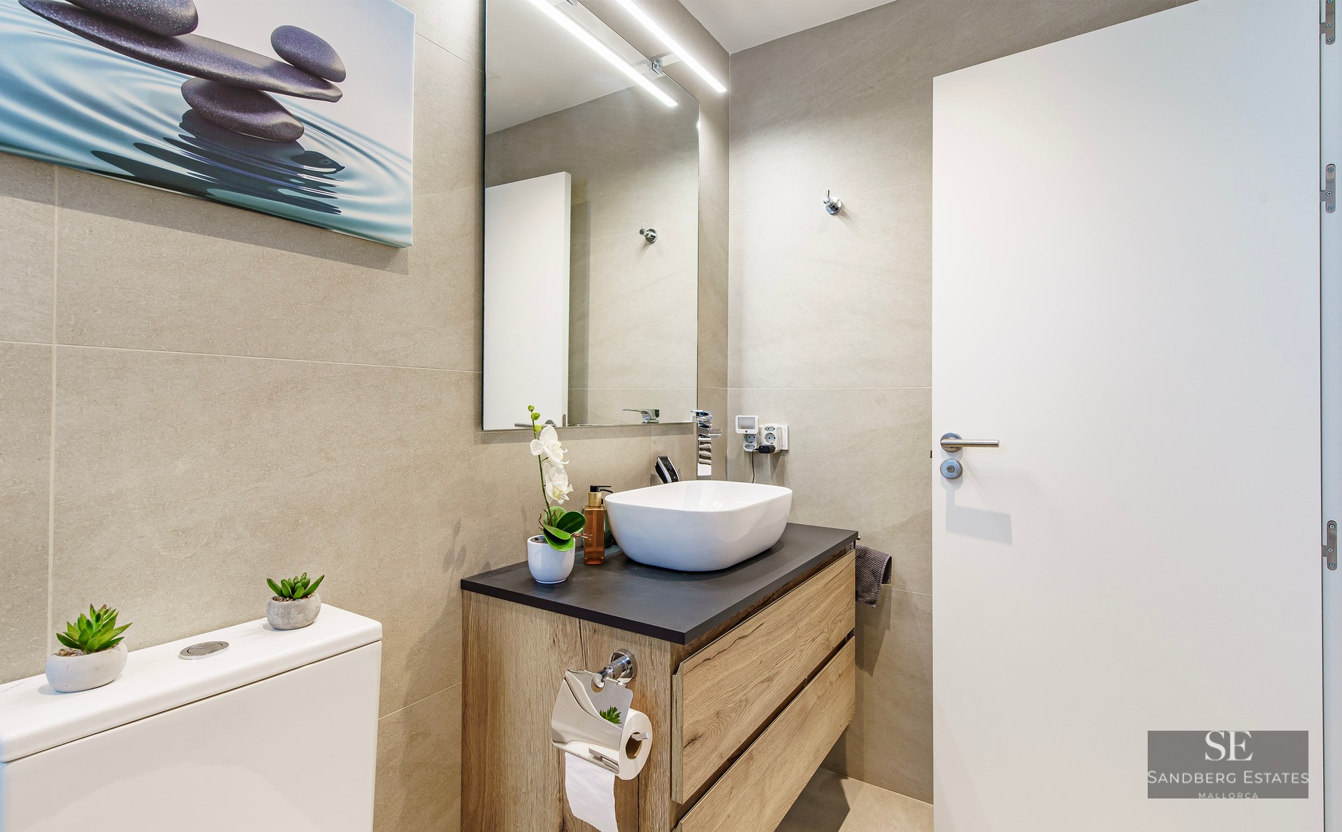 Modern bathroom featuring a white vessel sink on a wooden vanity, beige tiled walls, and a large mirror with LED light.