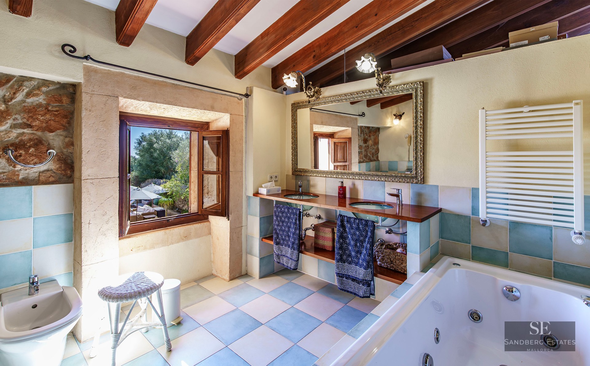 Master bathroom with exposed wood beams, double glass sinks, ornate mirror, and whirlpool tub overlooking a garden.