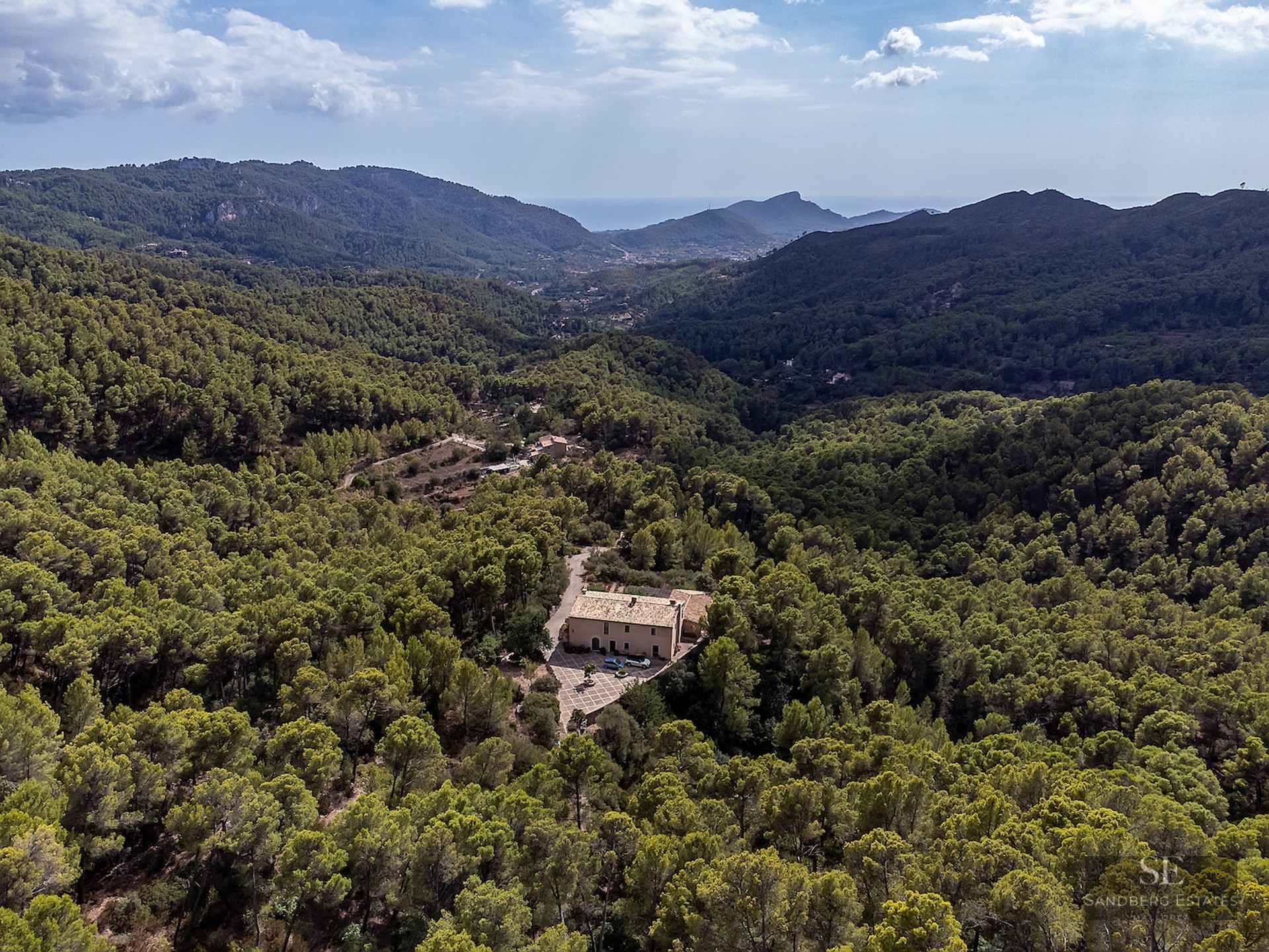 Aerial view of a private villa surrounded by dense green forest in a mountainous valley with a distant sea view.