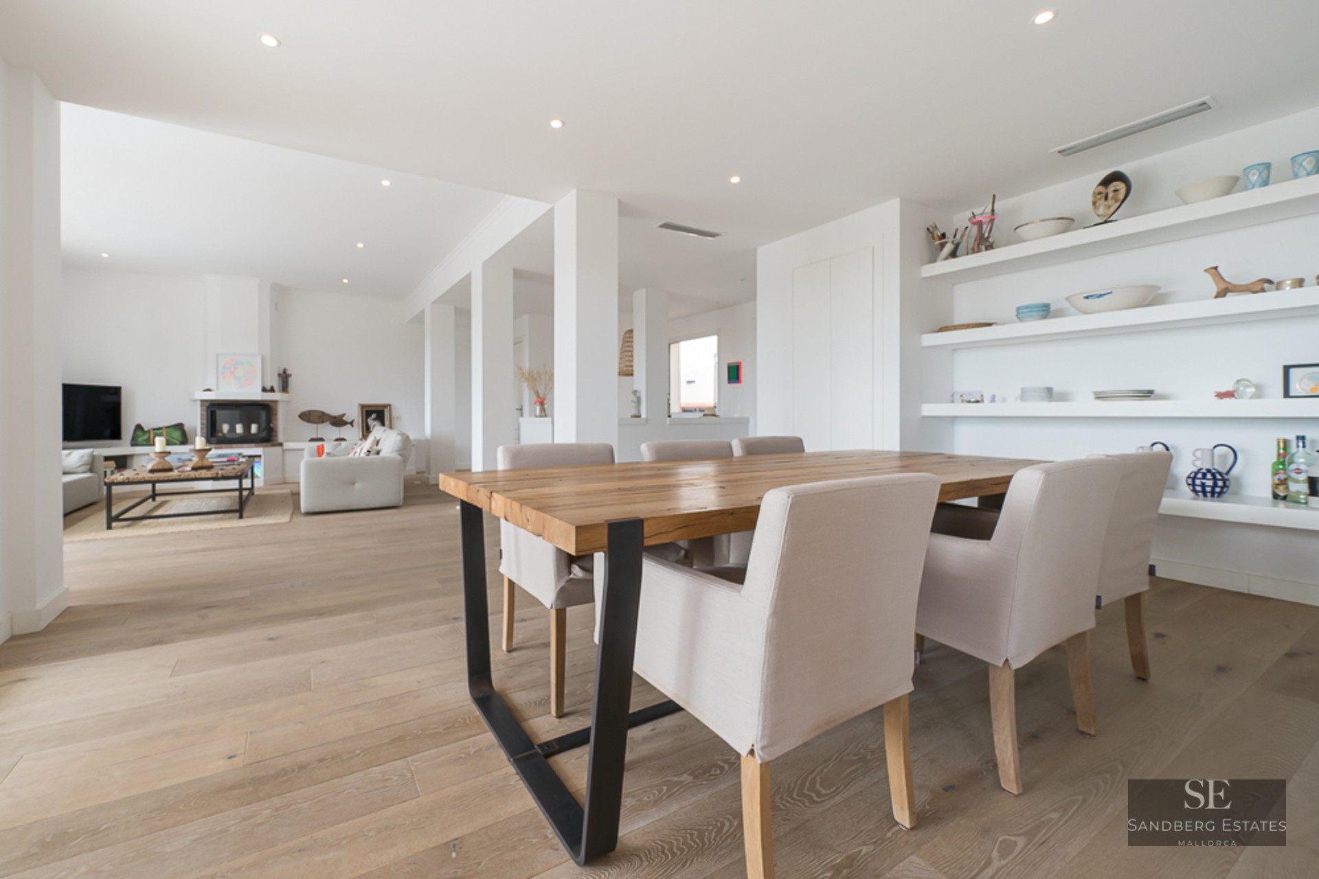 Salle à manger moderne avec grande table en bois et chaises rembourrées, s'ouvrant sur un salon lumineux avec cheminée.