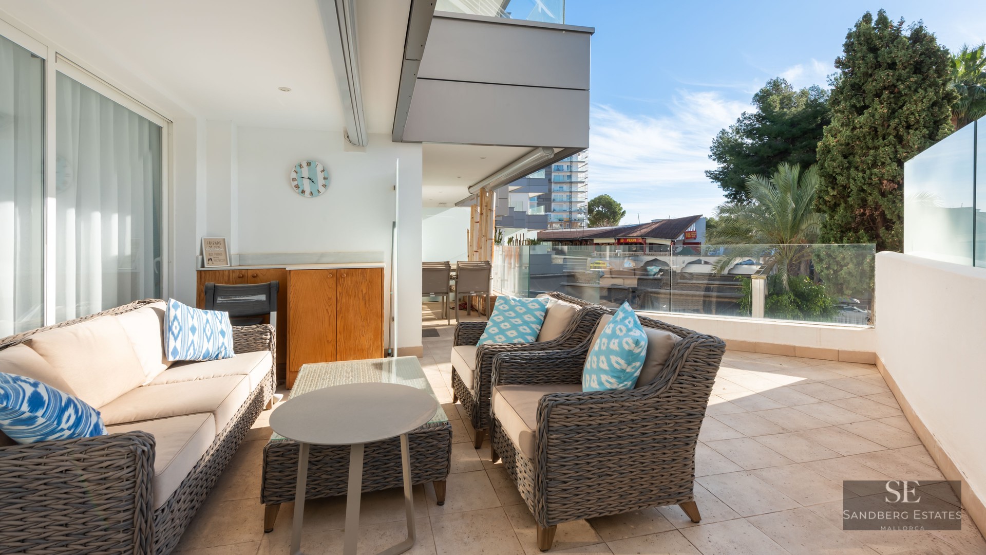 Grande terrasse avec canapés en osier gris, coussins bleus et garde-corps en verre sous un ciel bleu.