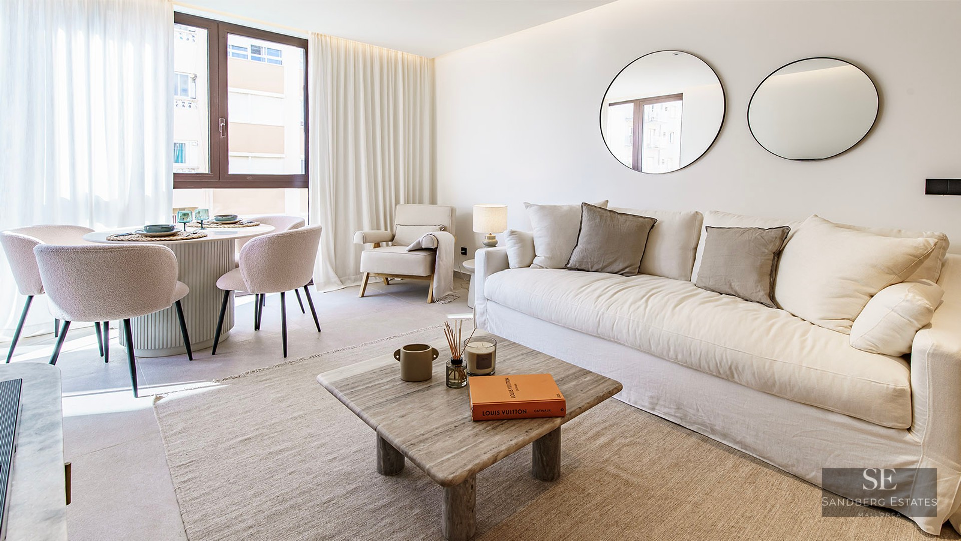 Bright living room featuring a large white sofa, stone coffee table, two round mirrors, and a dining table by a window.