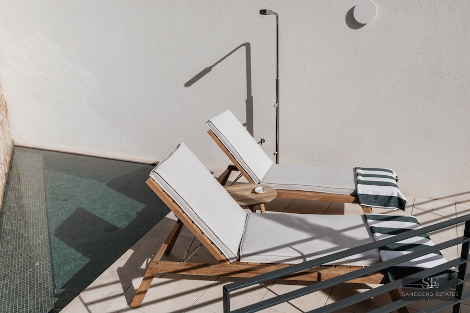Two wooden sun loungers with white cushions next to a pool and outdoor shower on a sunny terrace.