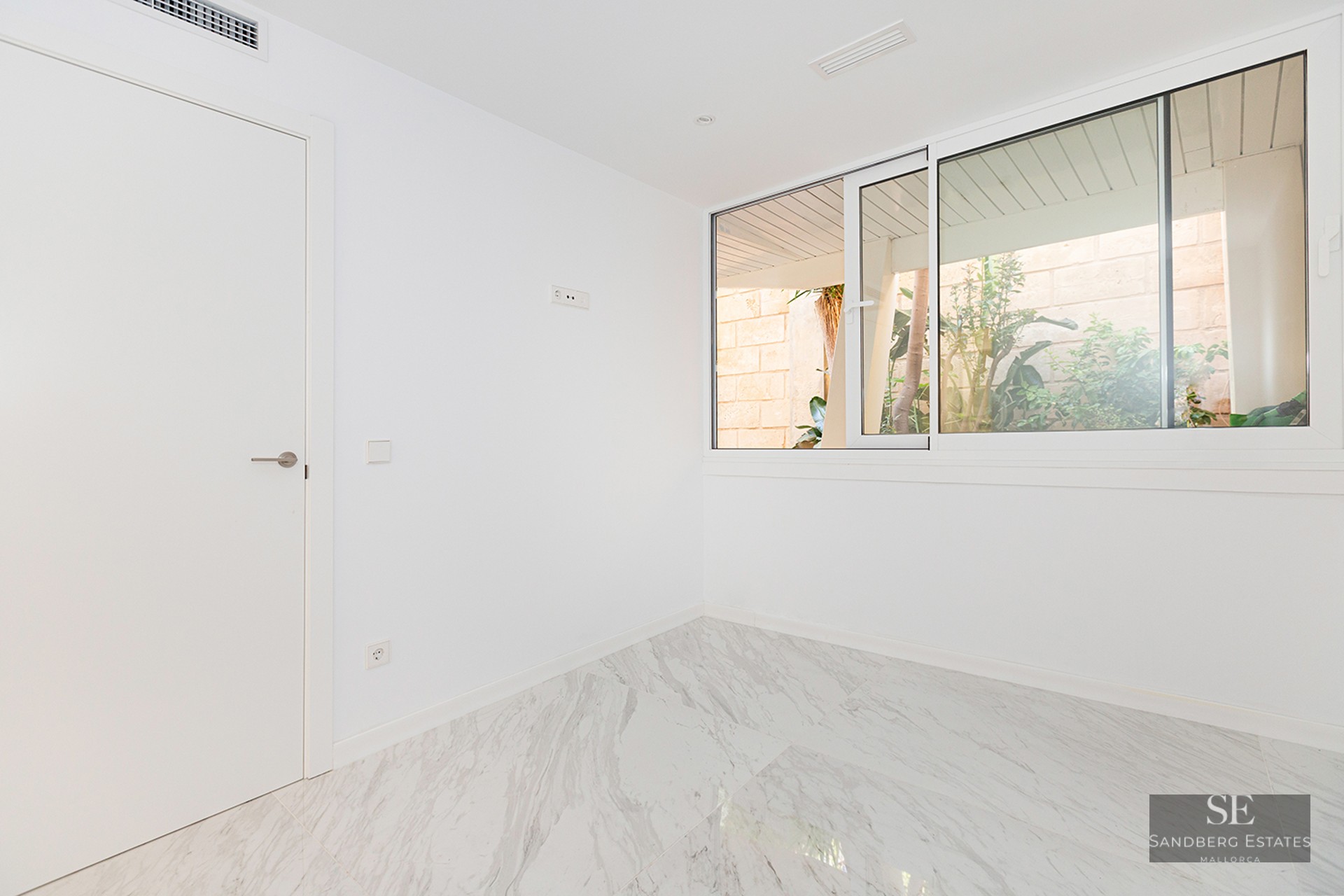 Empty room with white walls, polished marble floor, and a large sliding window overlooking outdoor greenery.
