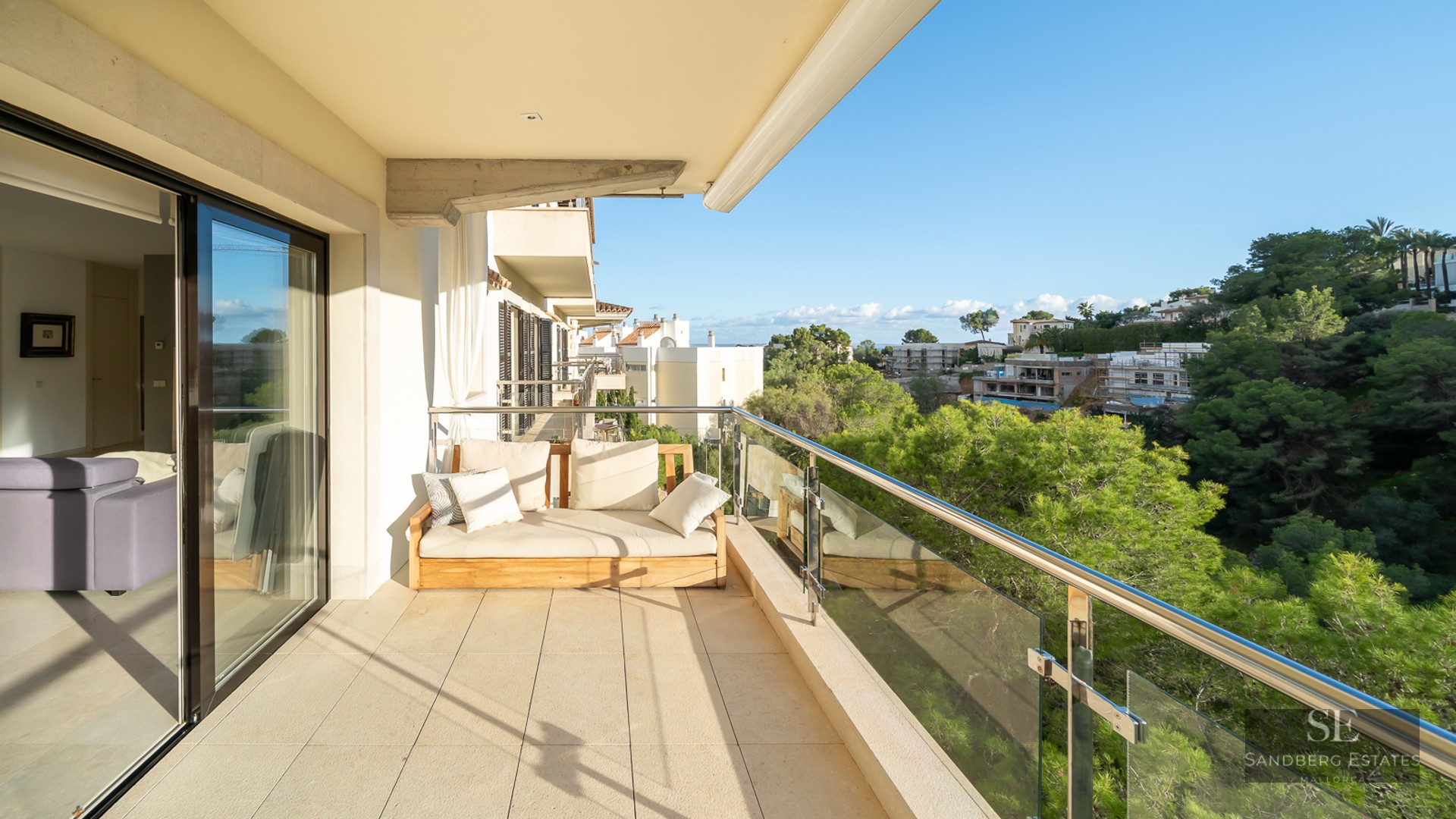 Modern balcony with wooden sofa and glass railing overlooking a lush green pine forest under a clear blue sky.