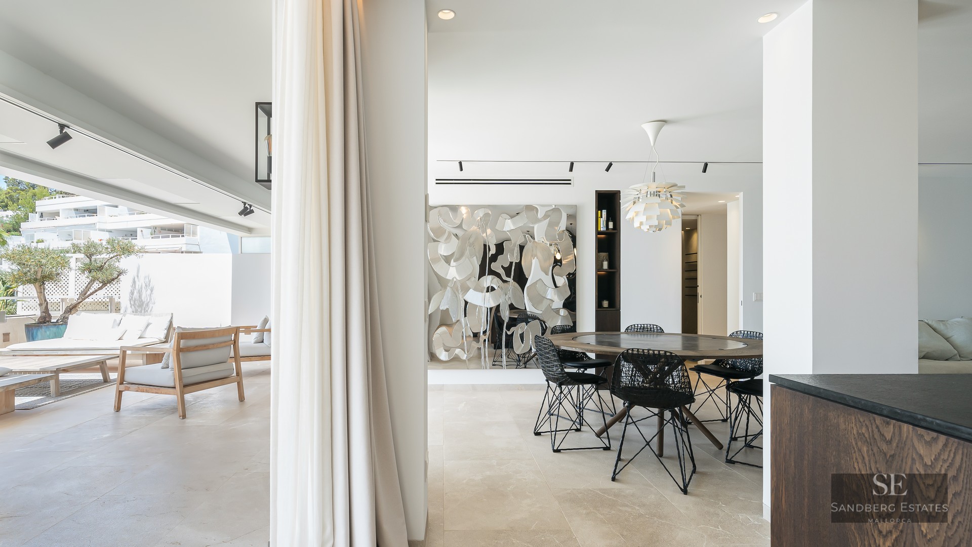 Modern indoor dining area with round wooden table opening onto a bright sun-drenched terrace with lounge seating.