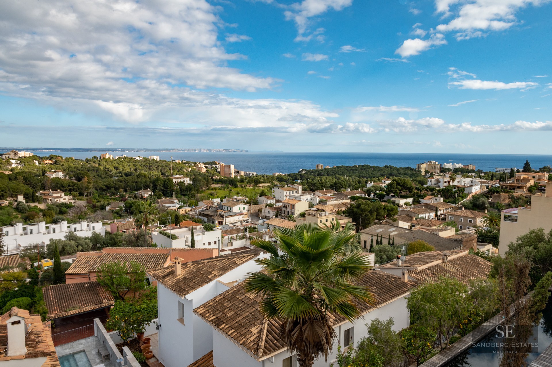 Wide angle view of white villas with terracotta roofs overlooking the blue sea under a partly cloudy sky.