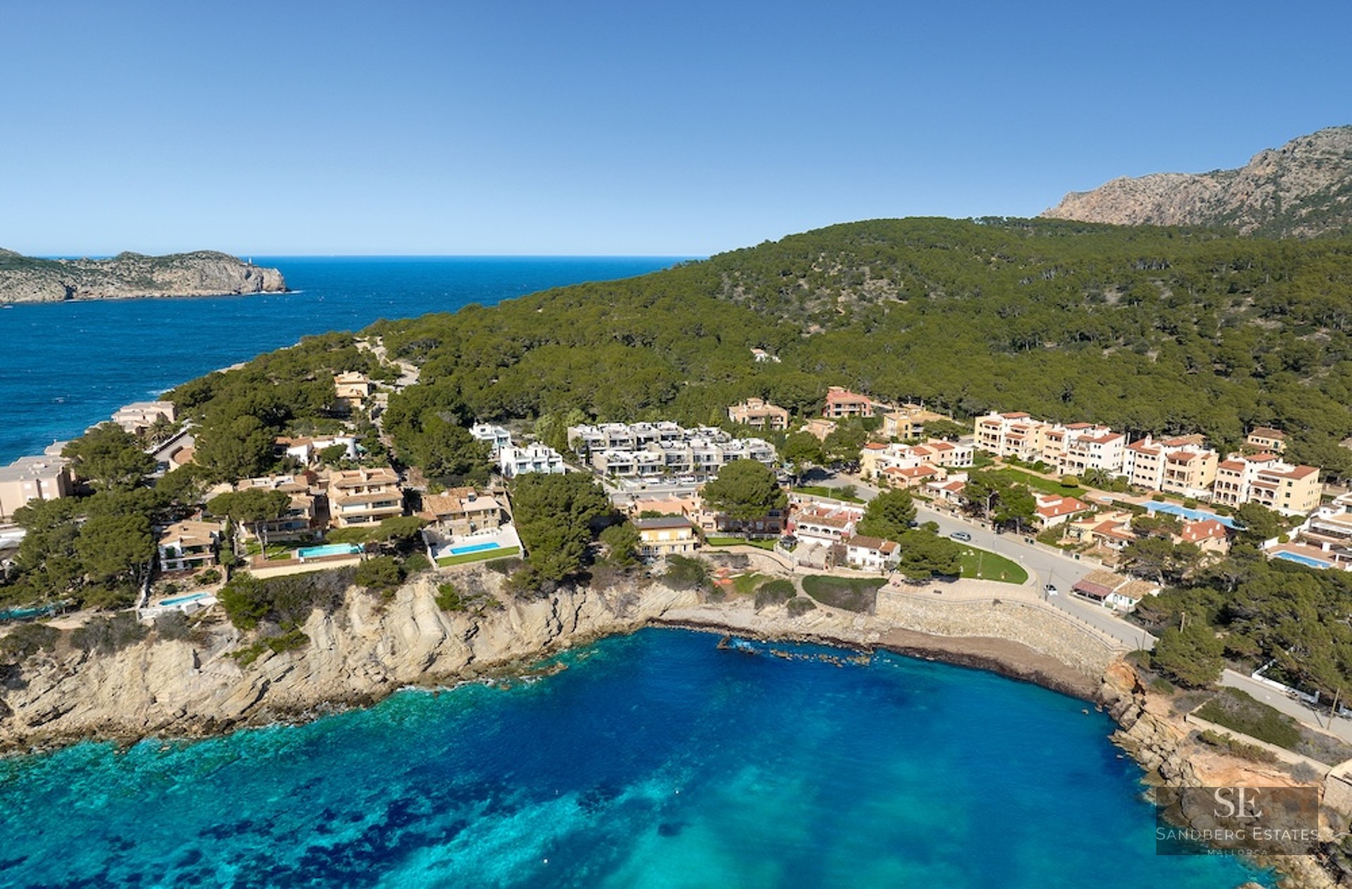 Aerial shot of Mediterranean villas on a rocky cliffside overlooking a turquoise bay and lush green forest.