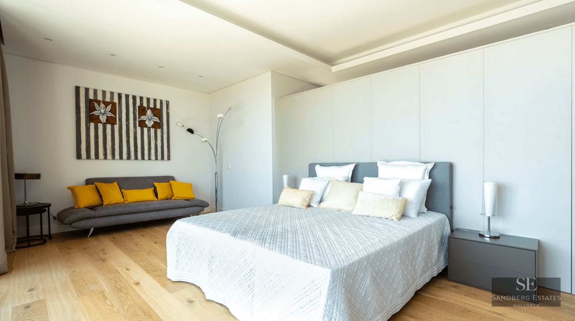 Bright modern bedroom featuring a large bed, wooden floors, and a grey sofa with yellow pillows.