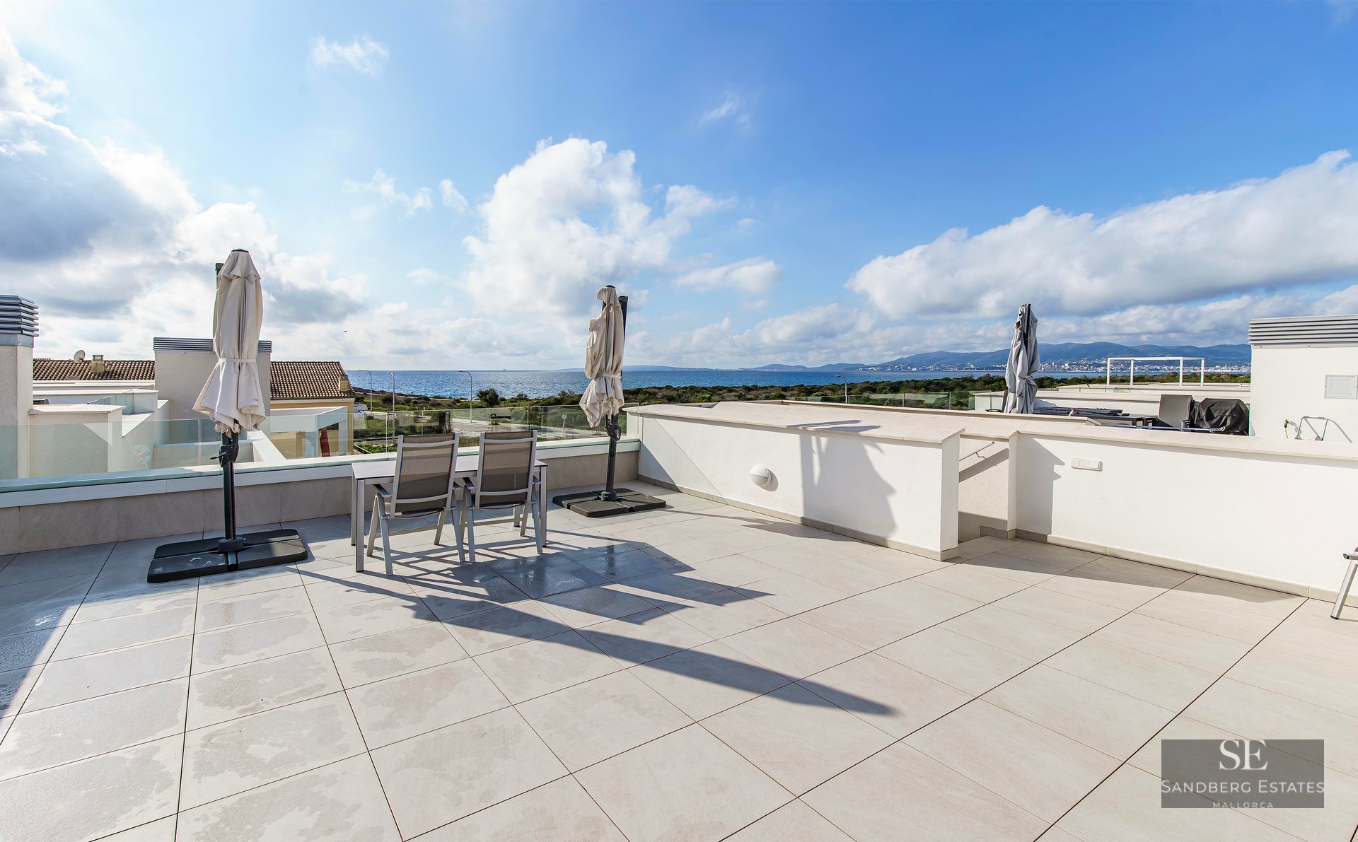 A large modern rooftop terrace with white tiles, glass railings, and a dining set overlooking the blue Mediterranean sea.