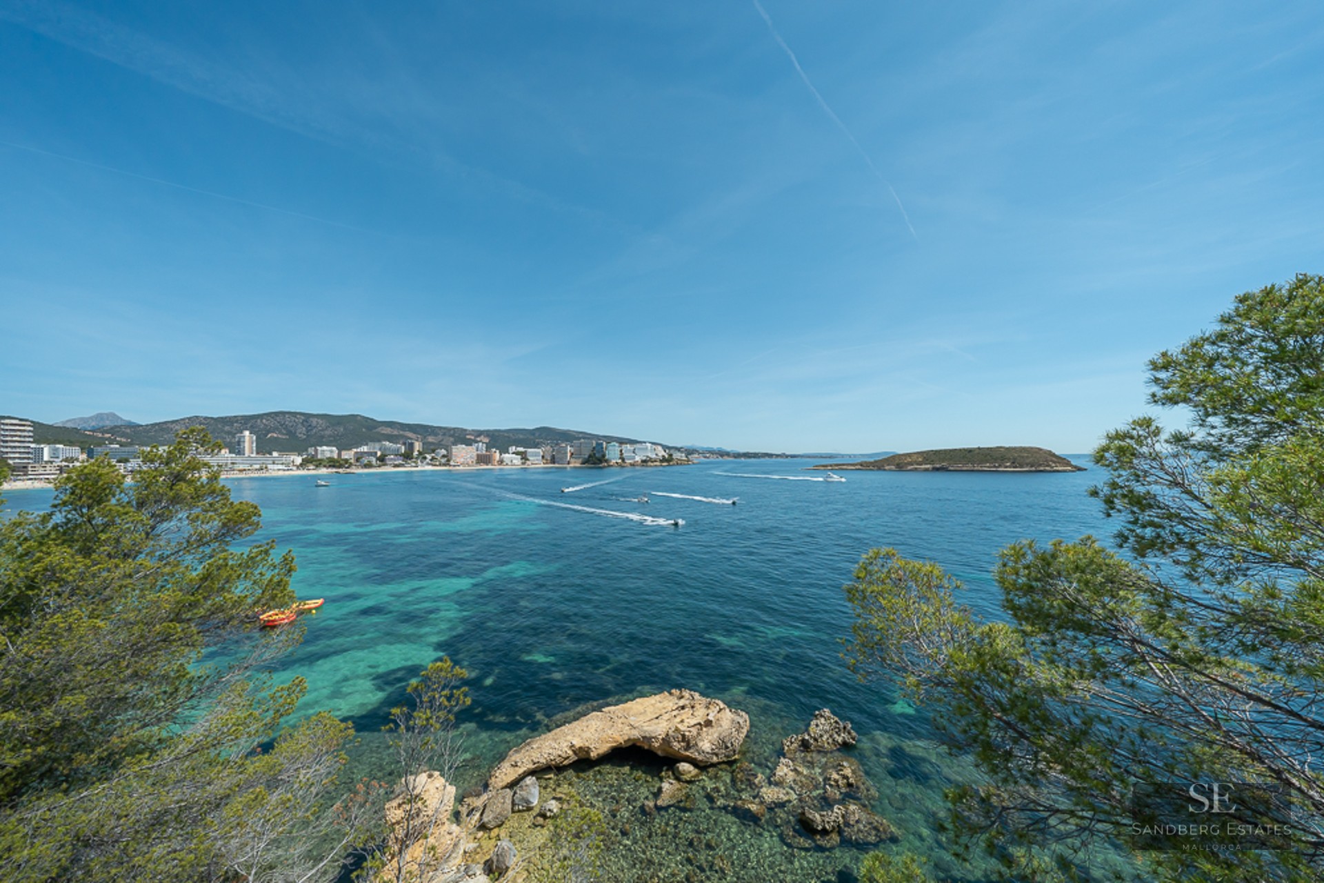View of a turquoise bay with boats, framed by pine trees and a coastal town in the background.