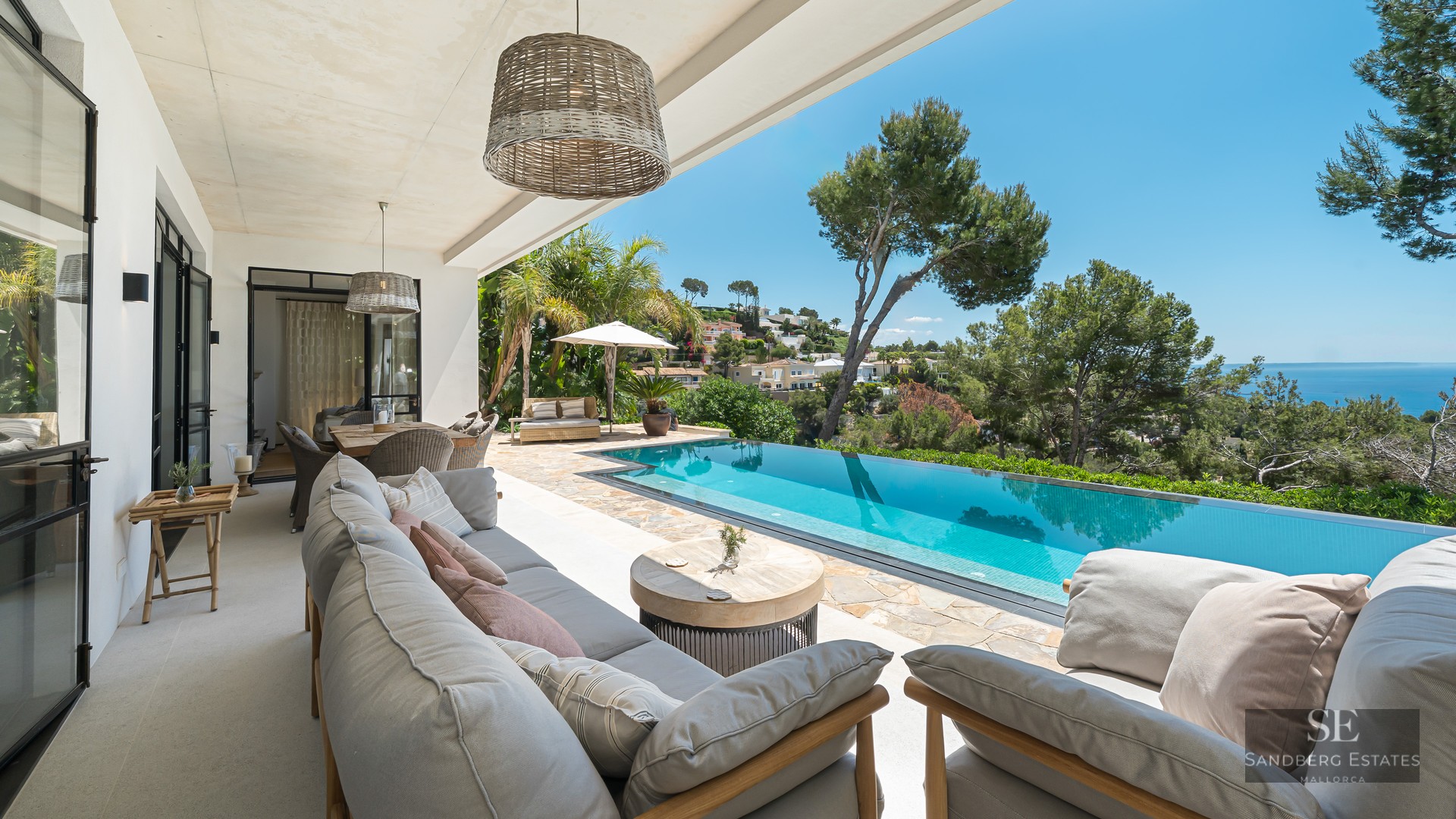 Covered terrace with grey lounge seating overlooking a turquoise infinity pool and the Mediterranean sea.