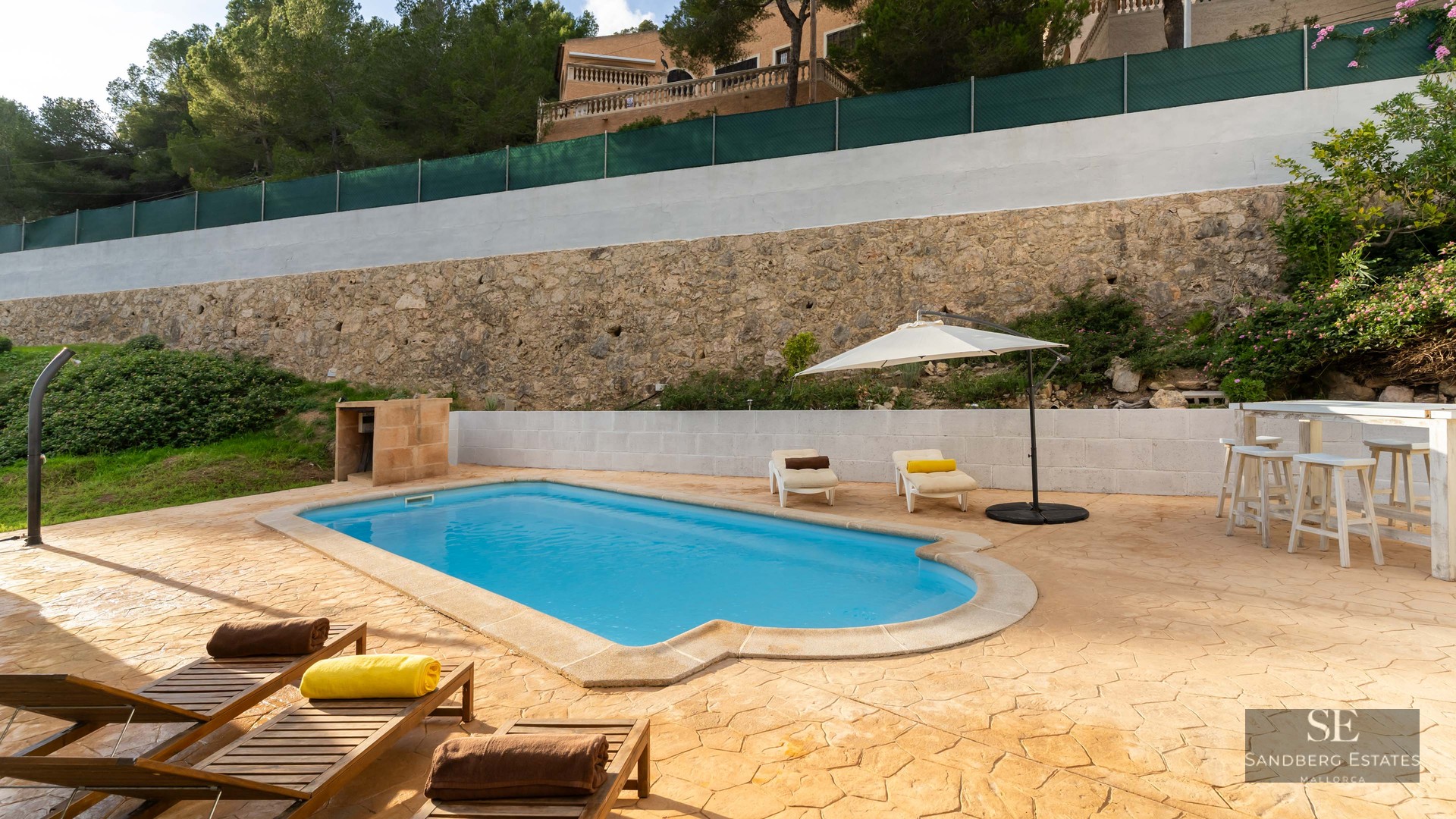 Piscine extérieure avec terrasse en pierre, chaises longues, parasol blanc et mur de pierre.