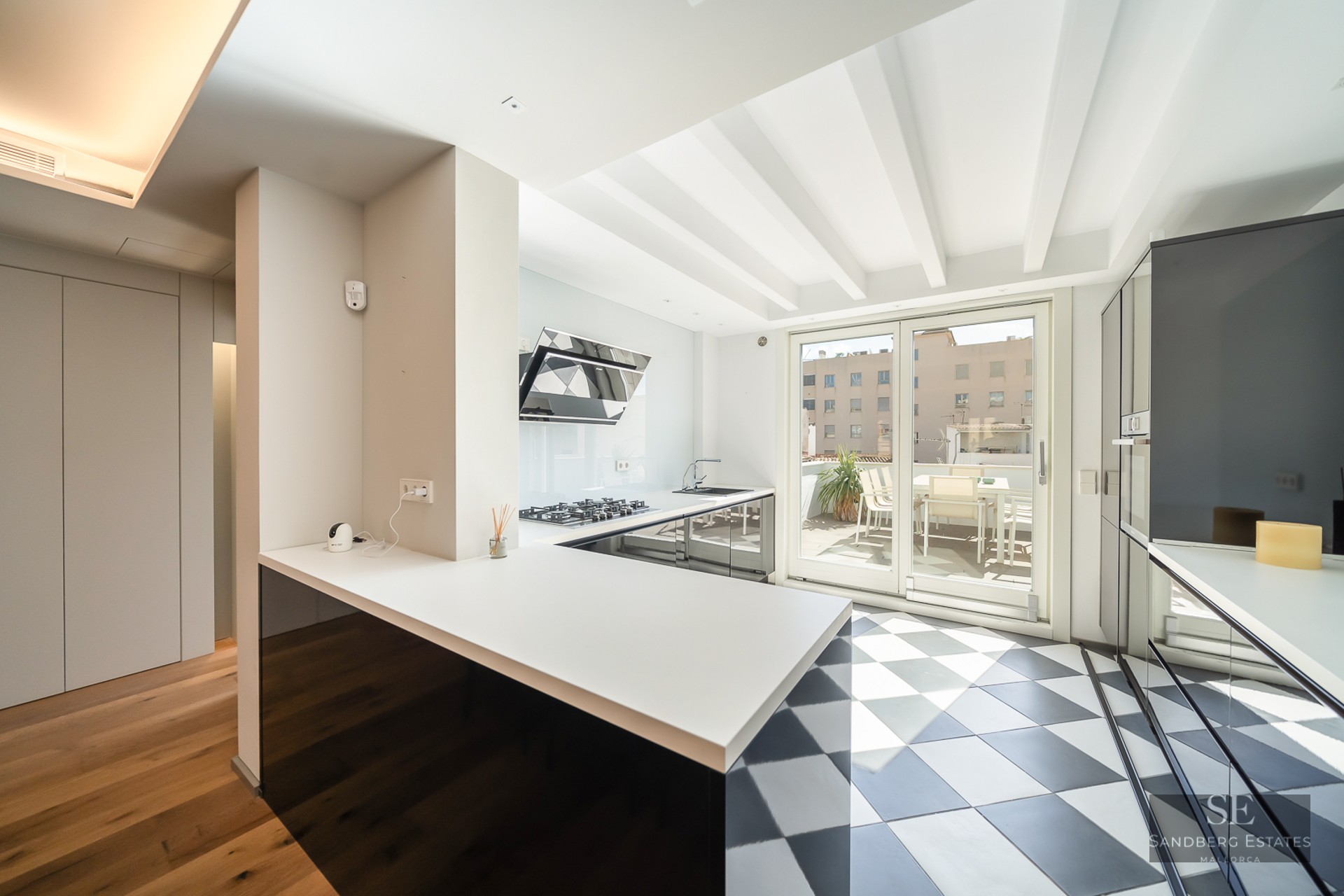 Modern kitchen featuring a black and white checkered floor, white ceiling beams, and large glass doors to a sunny terrace.