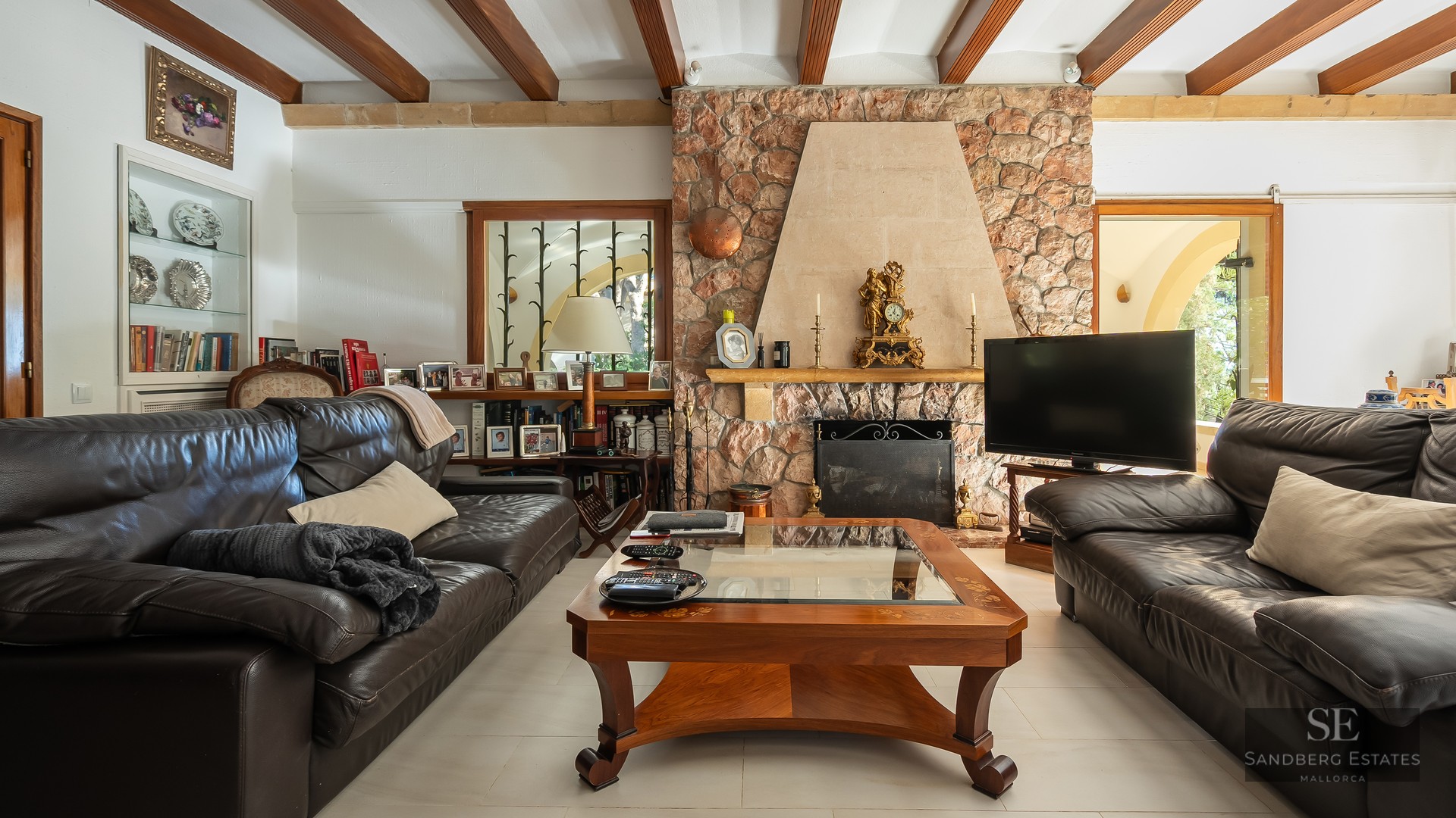 A cozy living room featuring dark leather sofas, a large stone fireplace, and exposed wooden ceiling beams.