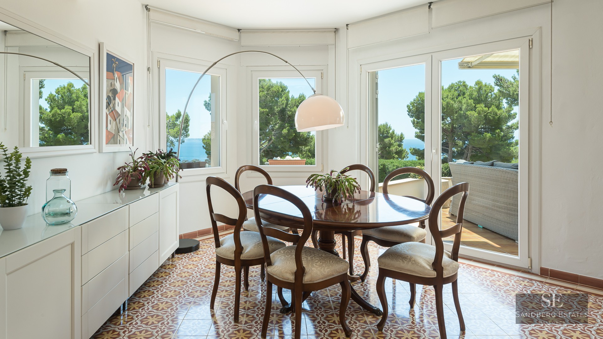 Bright dining room featuring a round wooden table, patterned tile floors, and large windows overlooking the sea.