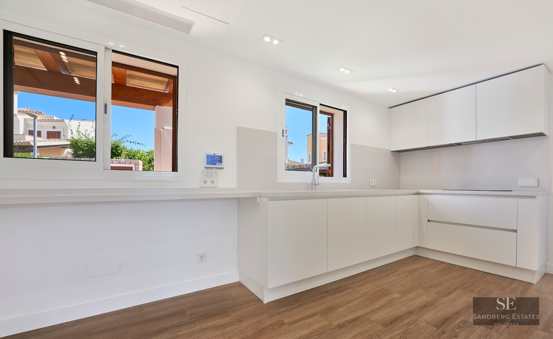 Modern white kitchen with handleless cabinets, wooden floors, and large windows with natural light.