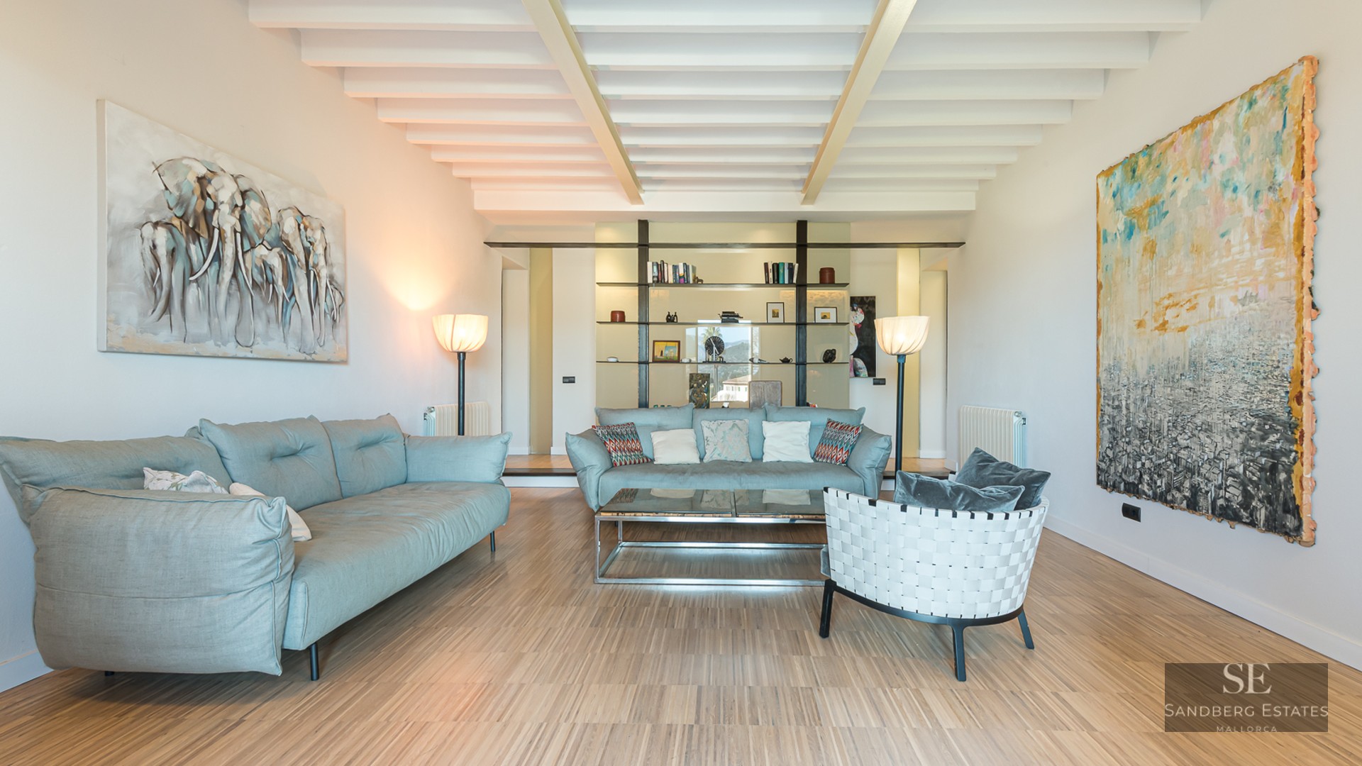 Spacious living room with light blue sofas, white beamed ceiling, wooden floors, and large abstract paintings.