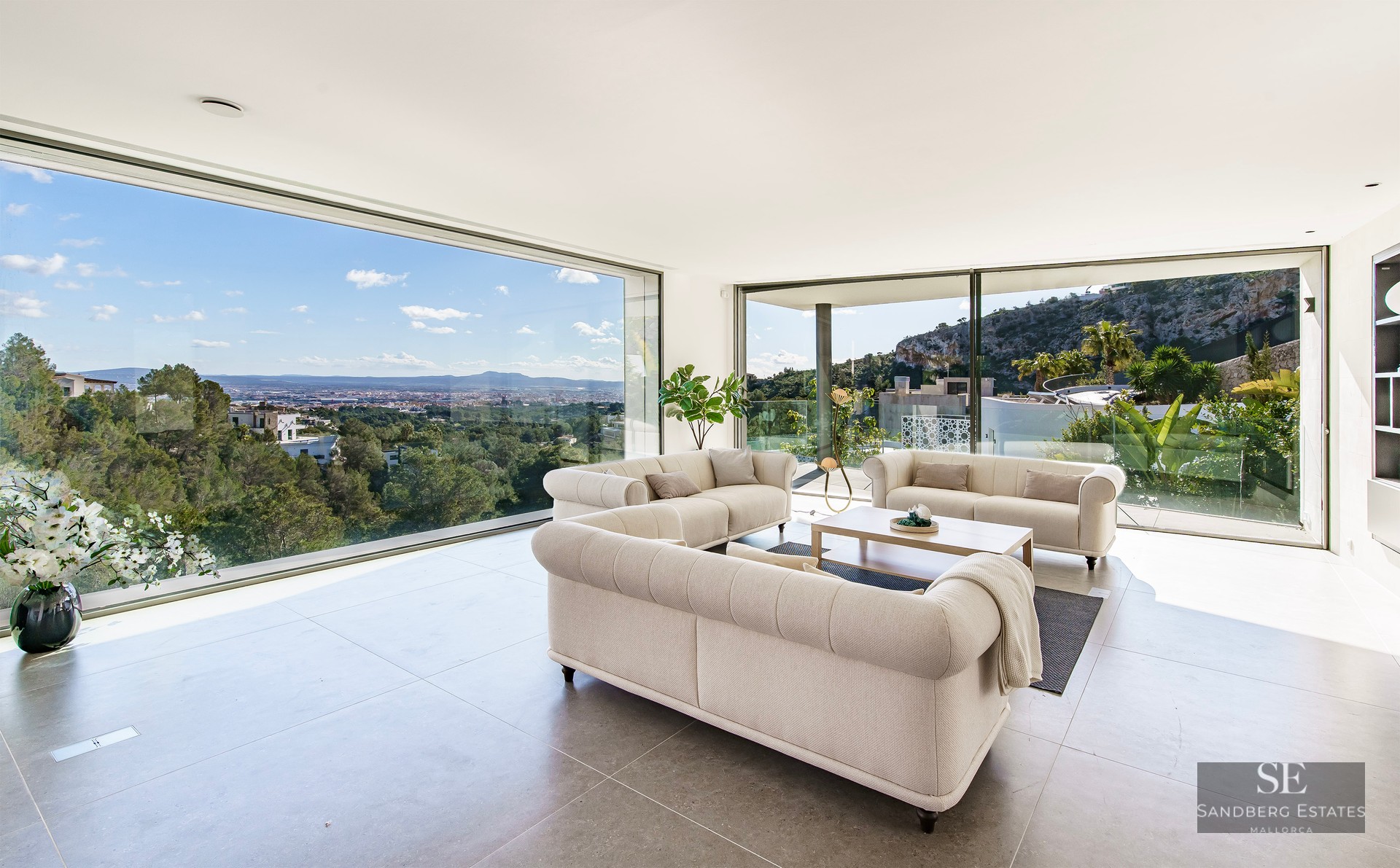 Modern living room with three beige tufted sofas and floor-to-ceiling windows overlooking a city valley and mountains.