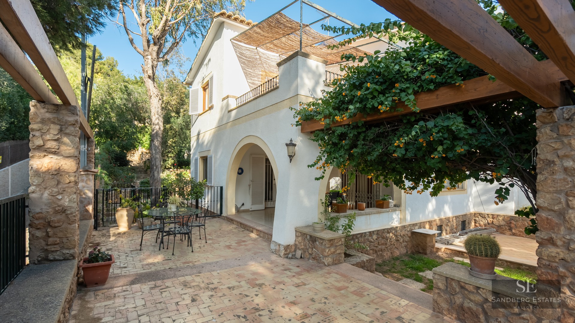 Sunny tiled patio with a flowering wooden pergola and white villa facade featuring arched entrance.
