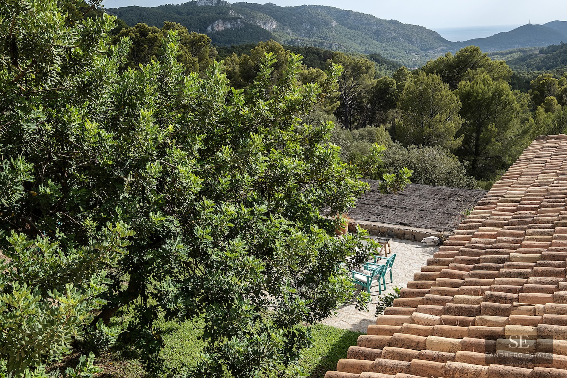 Terracotta roof overlooking a stone patio with teal chairs, surrounded by lush trees and distant mountains under a clear sky.